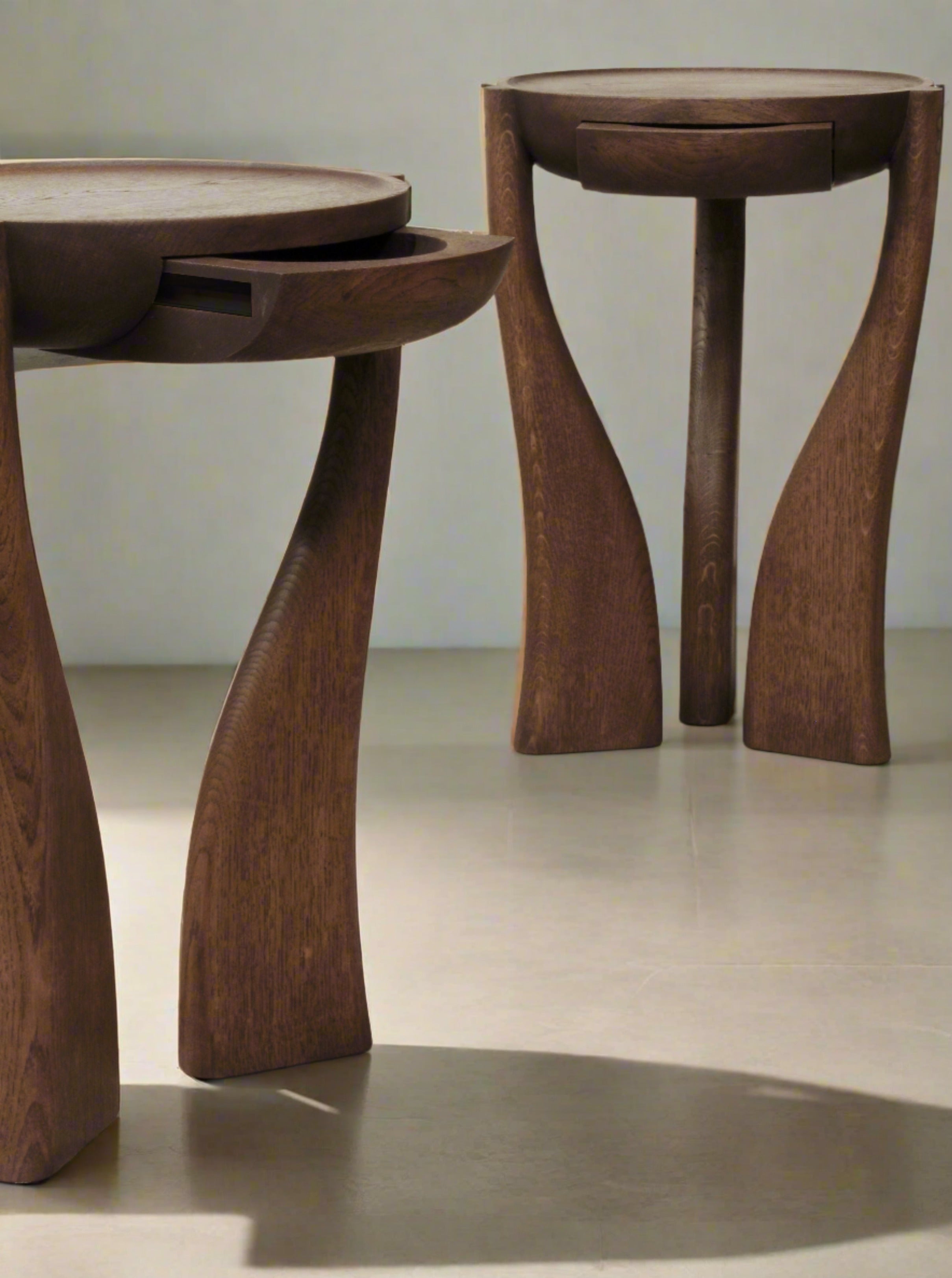 Two stylish Pair of End Tables UFØ - Limited Edition Exclusivity by Gaétan Le Gall with curved legs are shown in a well-lit room. Each table features a round top and a sleek, hidden drawer seamlessly integrated under the tabletop. These limited edition pieces are made from dark French oak and rest on a light-colored floor.