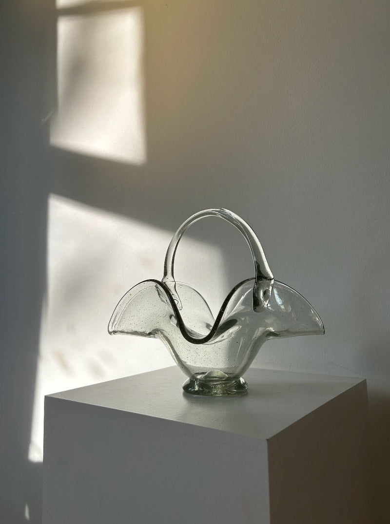 A 70's Vintage Glass Basket by Les Objets Amsterdam sits on a white pedestal against a plain wall. The basket, made of thick quality glass, has a unique, curvy design resembling an abstract flower with a handle. Bathed in soft, natural light and reminiscent of a 70's vintage bonbonnière, it creates a serene and minimalist scene. Shadows from a window pane fall gently on the wall.