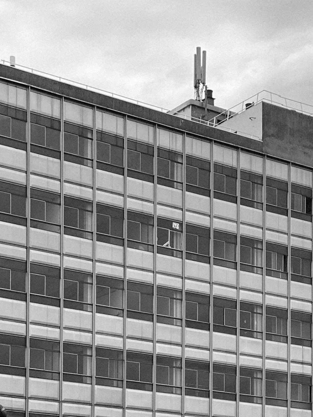 A black and white image captures a building facade echoing Bicci de' Medici's Jean Prouvé - Façade Panel 108, with rows of rectangular windows, some blinds partially drawn. A cell tower tops the roof, surrounded by silhouetted structures against a cloudy sky.