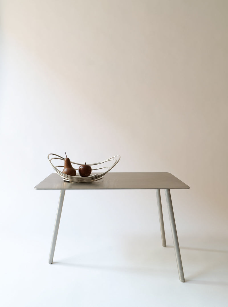 A simple, modern table with a smooth surface and four angled legs stands against a plain background. On top of the table is a decorative bowl containing two pears, serving as a Centrepiece Lino Sabattini 1970s from Collection apart. The overall aesthetic is minimalist and clean.