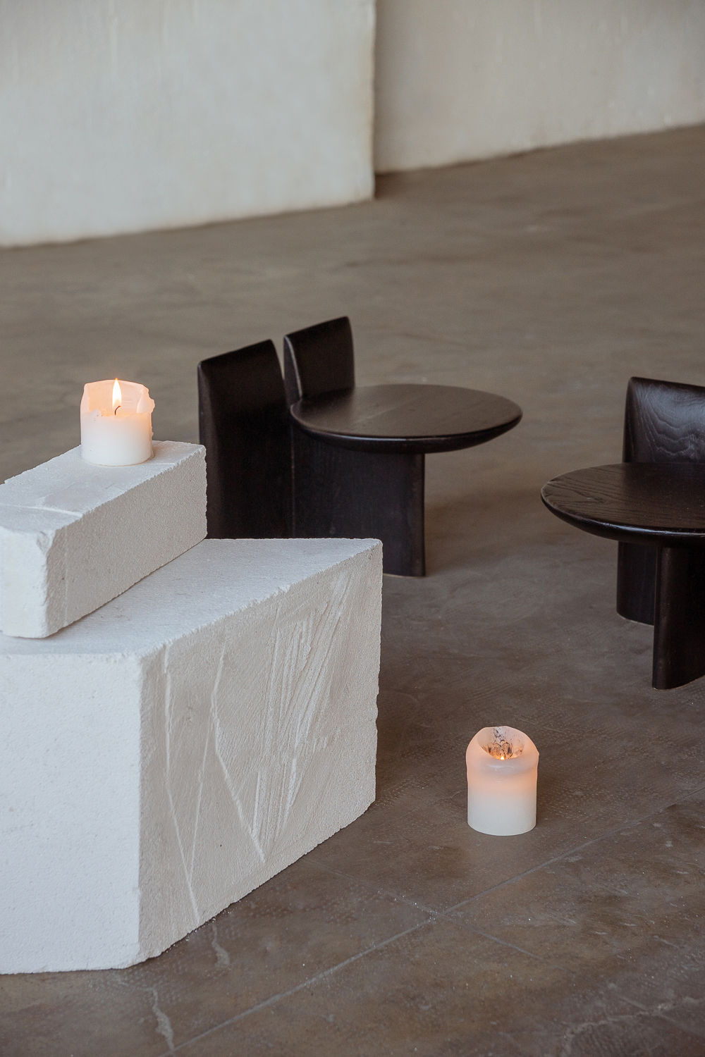 Two lit candles sit on white, textured blocks beside two black minimalist chairs and tables on a concrete floor. The setting features the Tabi Stand by Obstacles, using natural materials to exude modern serenity and artistic flair with soft lighting and sculptural stands.