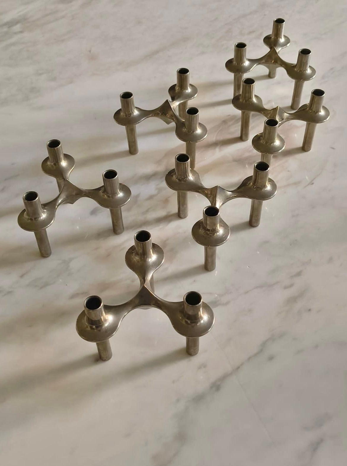 A collection of Rare Franz Nagel Candlesticks by Médecine arranged on a marble surface. These modular candle holders are interconnected, each featuring three slots to hold candles, forming intricate geometric formations. The midcentury steel candlesticks boast a shiny silver finish that reflects light beautifully.