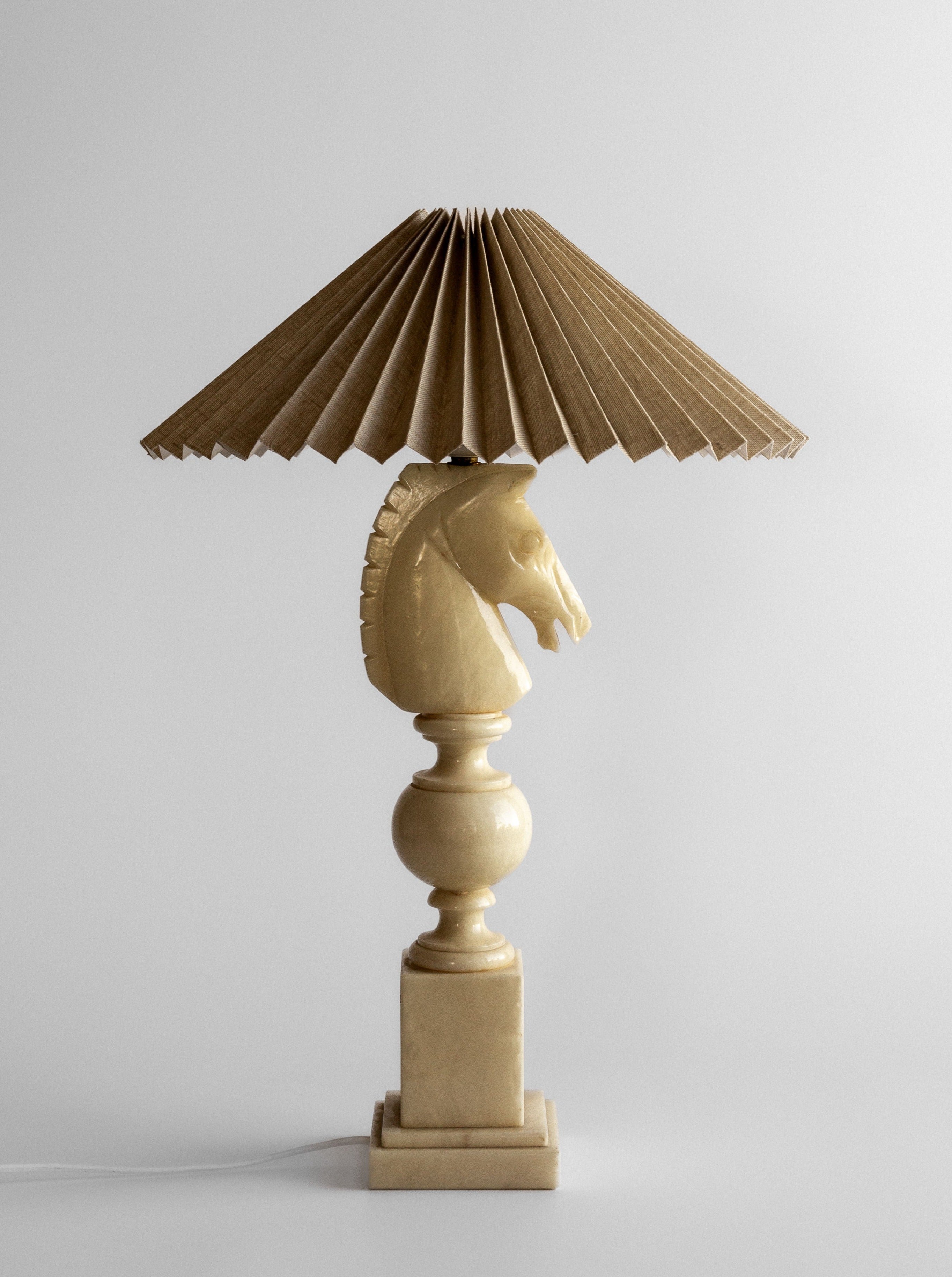 The White Knight Alabaster Table Lamp by Spigel, crafted in Italy during the 1970s, features a distinct horse head sculpture reminiscent of a chess piece design, mounted on a square pedestal. The lampshade, made of pleated beige fabric resembling a fan, adds texture and charm while emitting a warm and inviting glow.