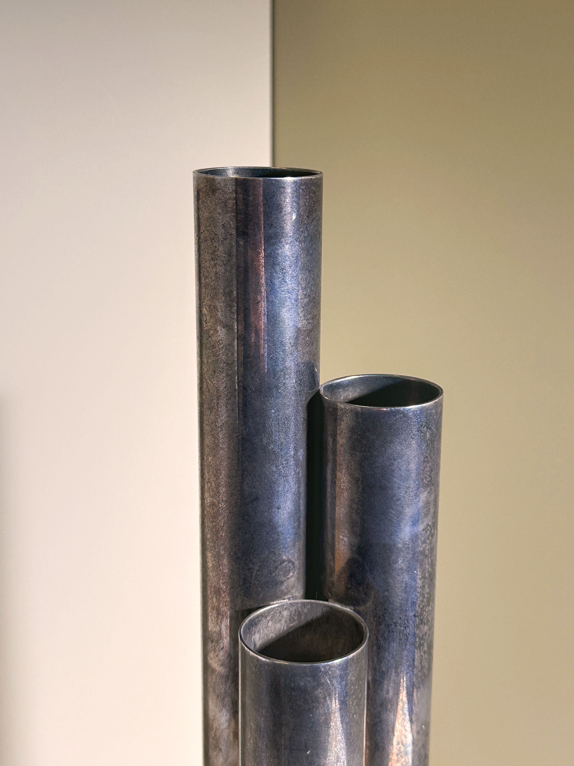 A set of four metallic pipes, designed as the Silver-plated Tall Brutalist Vase by Dodo Vintage, evokes a minimalist and industrial aesthetic reminiscent of Gio Ponti's 1960s designs against a neutral, shadowed backdrop.