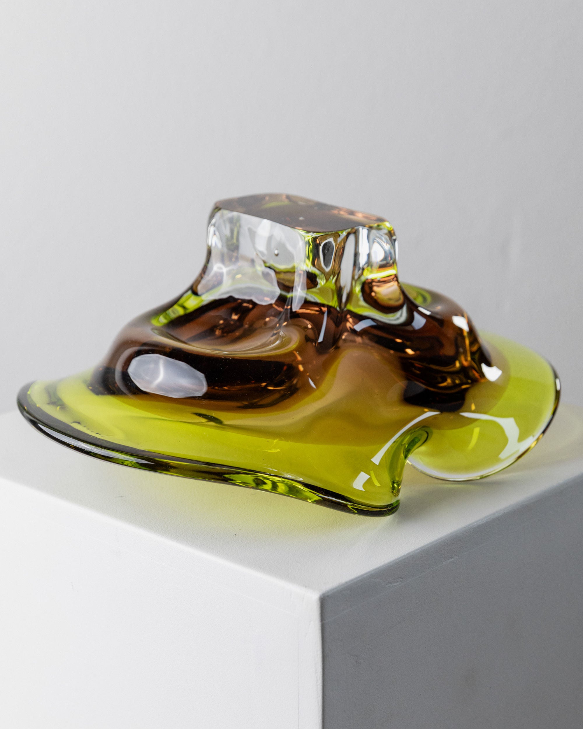 The Murano Glass Bowl by Bottega Jacobs, crafted in Italy in the 1970s, boasts a fluid, organic shape with translucent amber and green hues. This modern sculpture sits on a white pedestal and features a smooth, undulating surface reminiscent of melting forms with artistic flair.