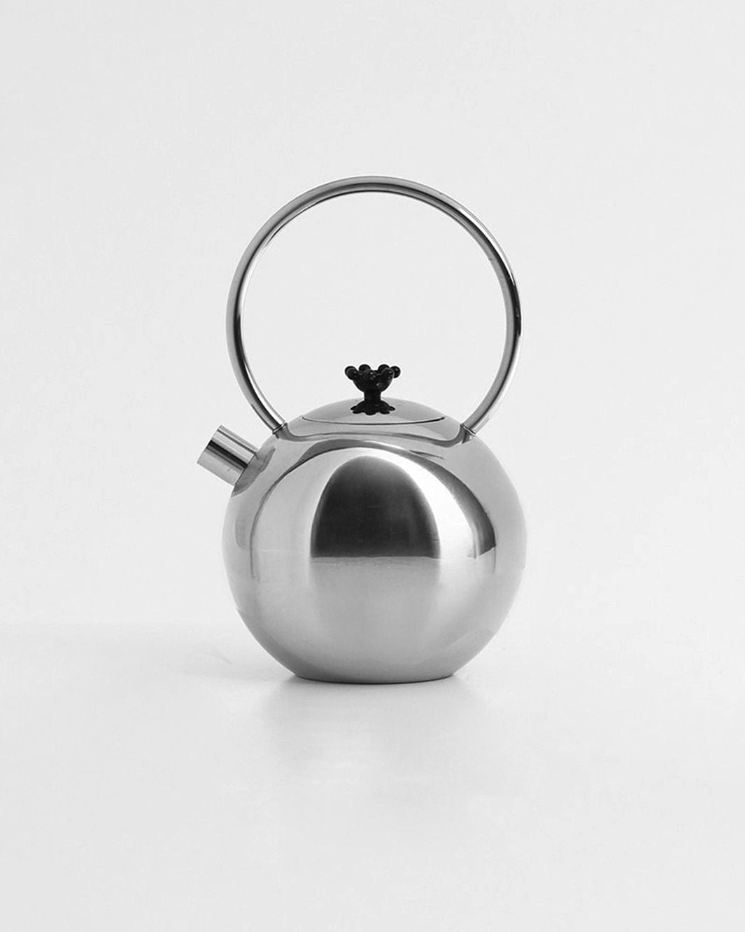 The Chrome teapot by Matteo Thun for WMF, available from Dodo Vintage, is a round and modern piece with a smooth stainless steel surface. It features a large circular handle on top and a short, angled spout. A small black knob adorns the lid, all contributing to its minimalist elegance against a plain white background.