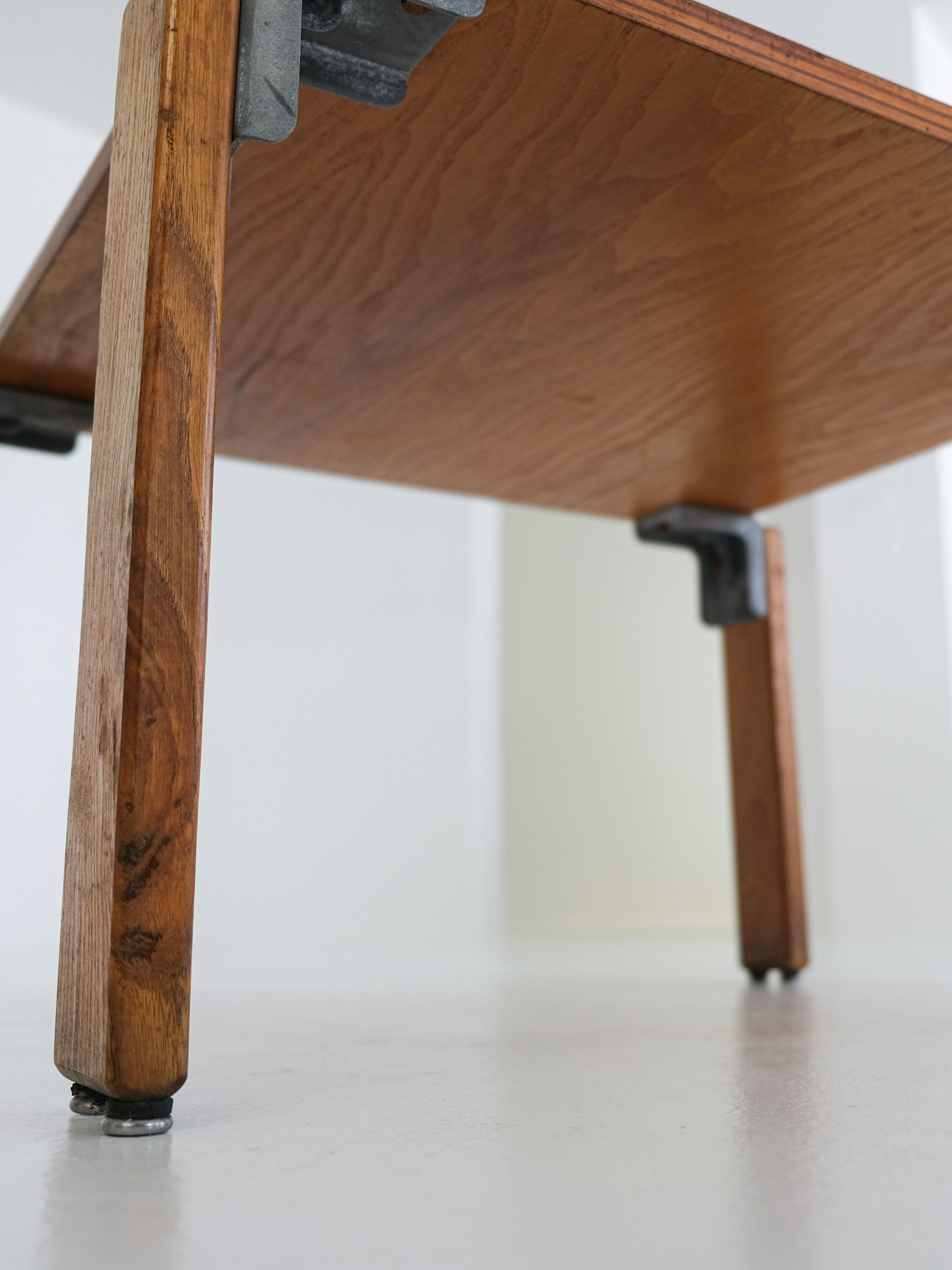 Close-up of the Coffee Table by George Candilis and Anja Blomstedt showcases its oak plywood legs with visible wood grain from below. Metal brackets connect the legs to the smooth, polished top, reflecting the simple 1970s design characteristic of antibeige.