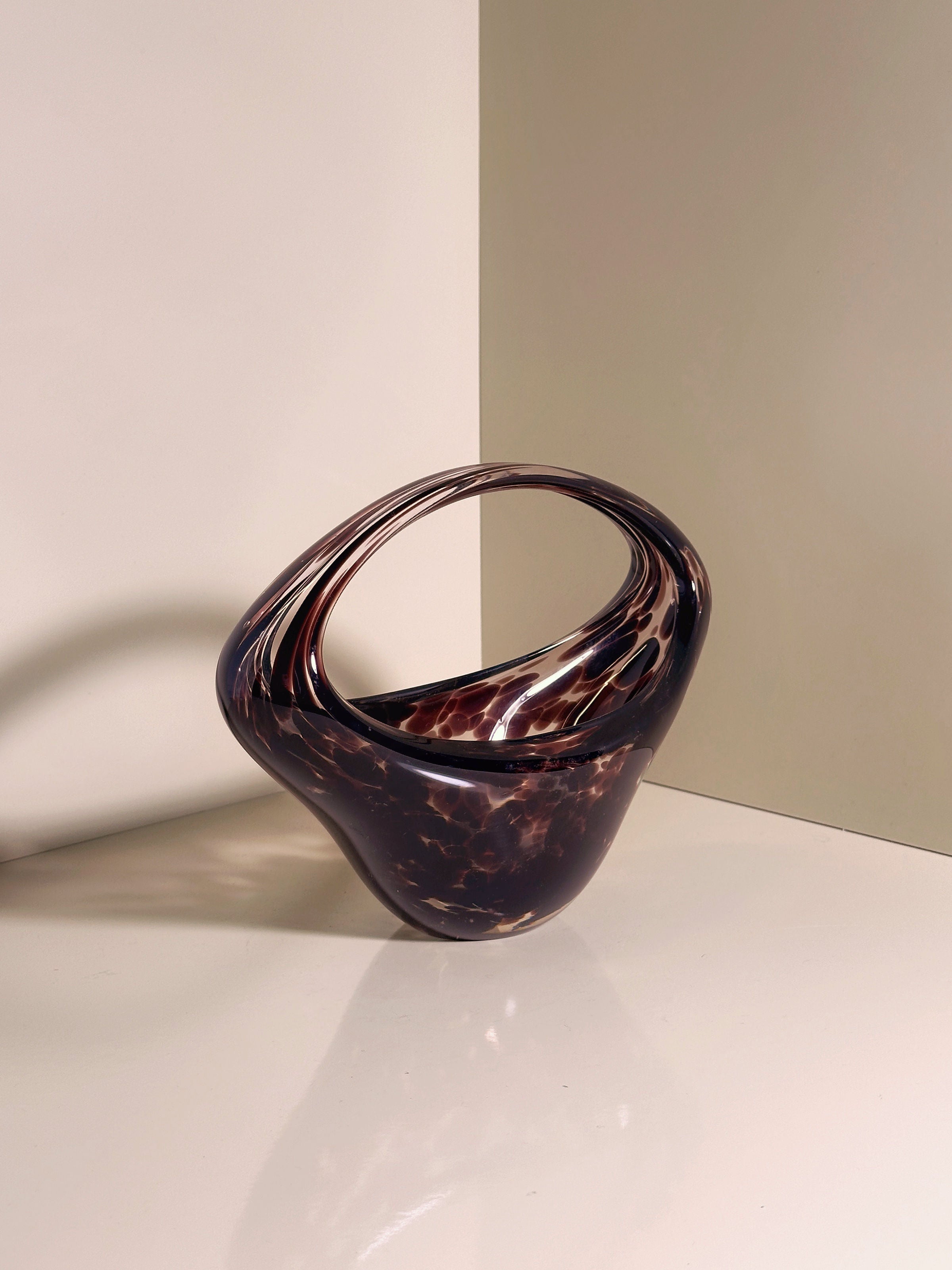 The Murano Tortoise Glass Handle Bowl from the 1970s by Dodo Vintage features a brown glass sculpture with a smooth, twisted loop design, casting subtle shadows on a glossy white surface against a soft two-toned beige wall, offering a sleek and modern aesthetic.