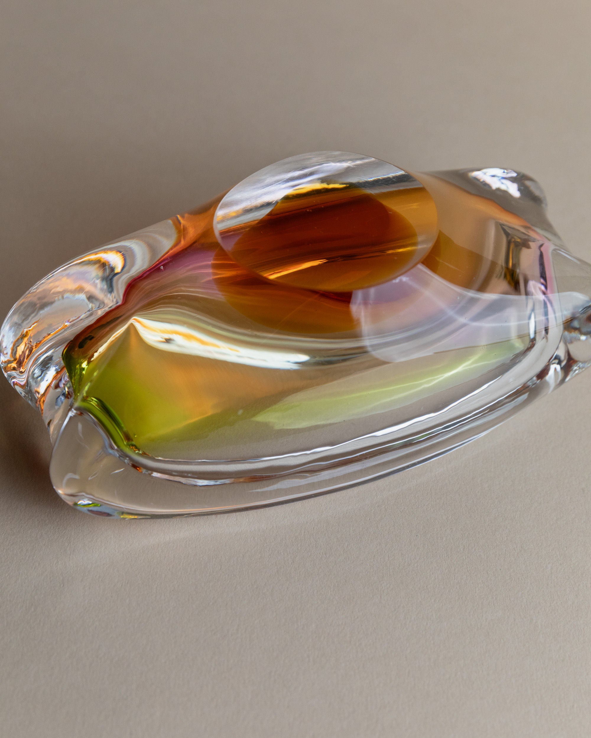 The Glass Ashtray by Josef Rozinek for Novy Bor, from Bottega Jacobs, features fluid hues of orange, yellow, and green on a neutral beige surface. Its smooth curves and oval indentation reflect light beautifully, mesmerizing onlookers with their soft shadows.