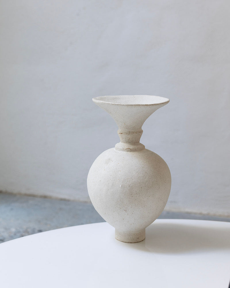 The Ánfora Blanco Hueso Vase by Canoa Lab, a white textured ceramic piece with a wide flared rim and spherical body, handmade in Valencia, rests on a smooth white surface against a plain light wall.