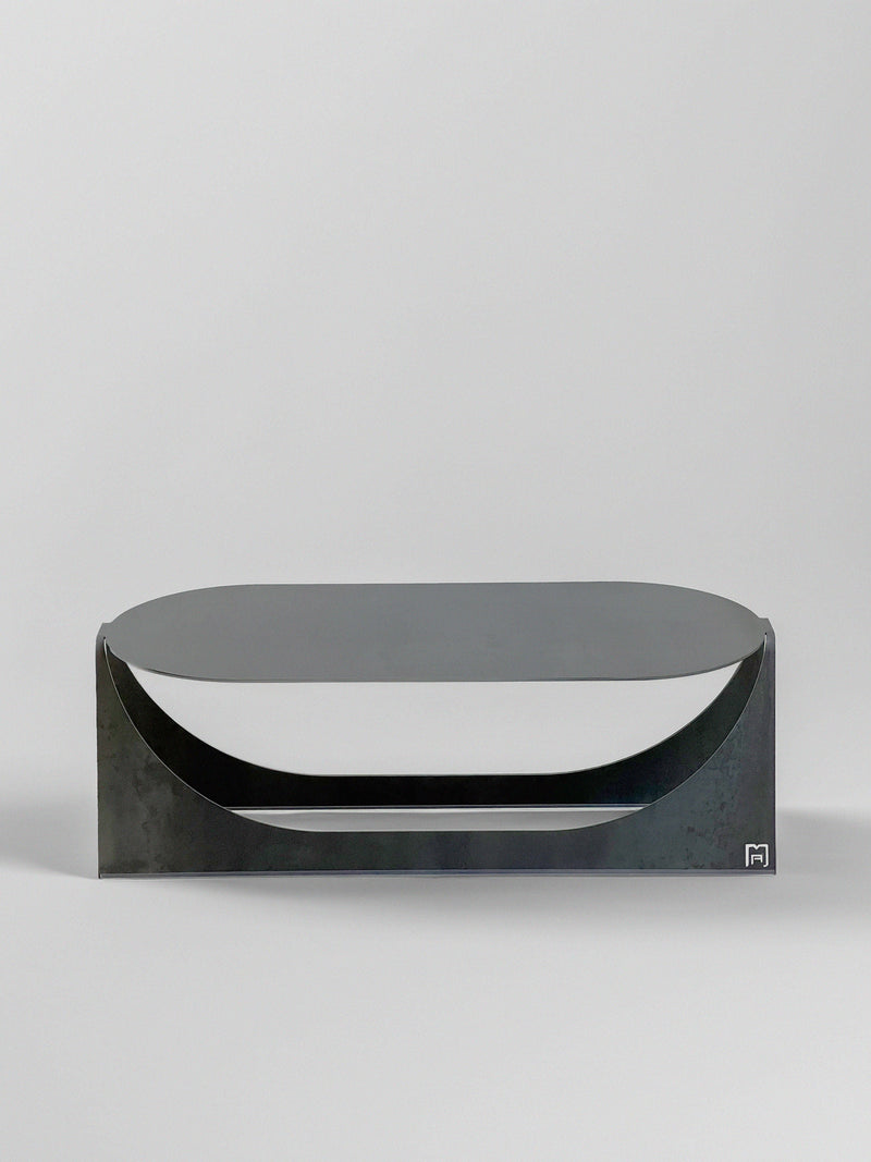 The 'Tani' Coffee Table by Modan is a modern, minimalist black metal piece with a flat top and curved cutout details on the front, crafted from steel plate, shown against a plain white background.