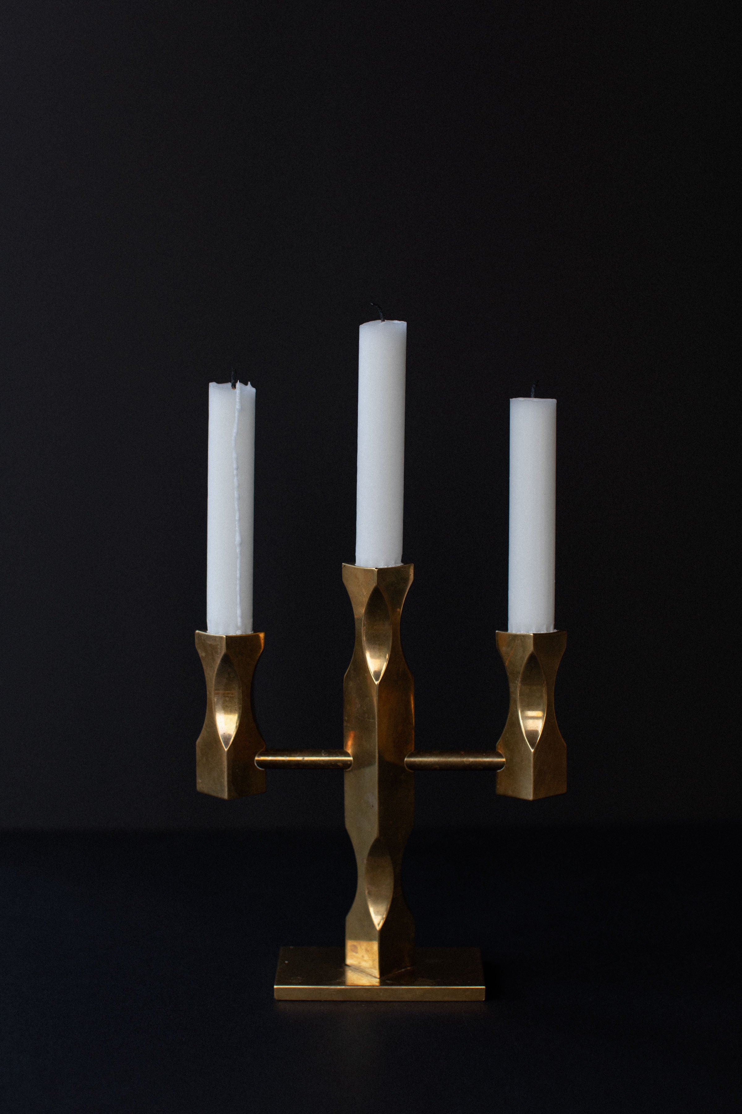 The Brass Candleholder by Out For Lunch, featuring a Brutalist design crafted from solid brass, stands against a dark background. Its angular and sculptural form holds three candles—two partially burned and the third yet to be lit.