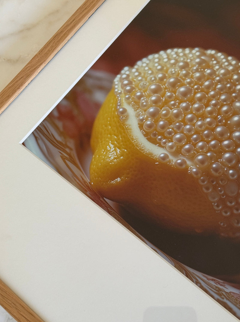 A framed fine art print of a lemon covered in shiny bubbles, likely water droplets, against a dark background. The light wooden border adds a touch of elegance and is placed on a light-colored surface, possibly marble. This mixed media photography piece, *Lemon and Pearls* by *Adeline Mai*, exudes sophistication.