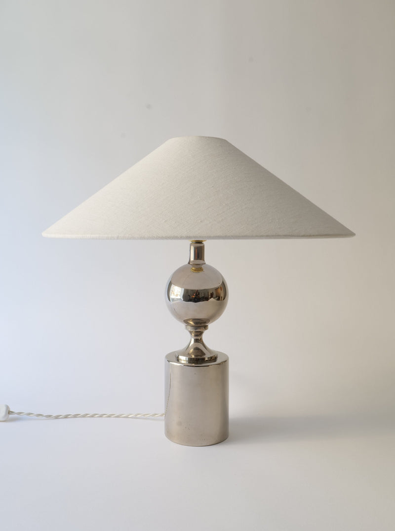 A Collection apart Barbier Lamp 1970s with a metallic cylindrical base and a spherical middle section, topped with a wide, cream-colored conical lampshade. Designed by Philippe Barbier, this nickel over brass table lamp features a white power cord attached at the bottom. The background is plain white.