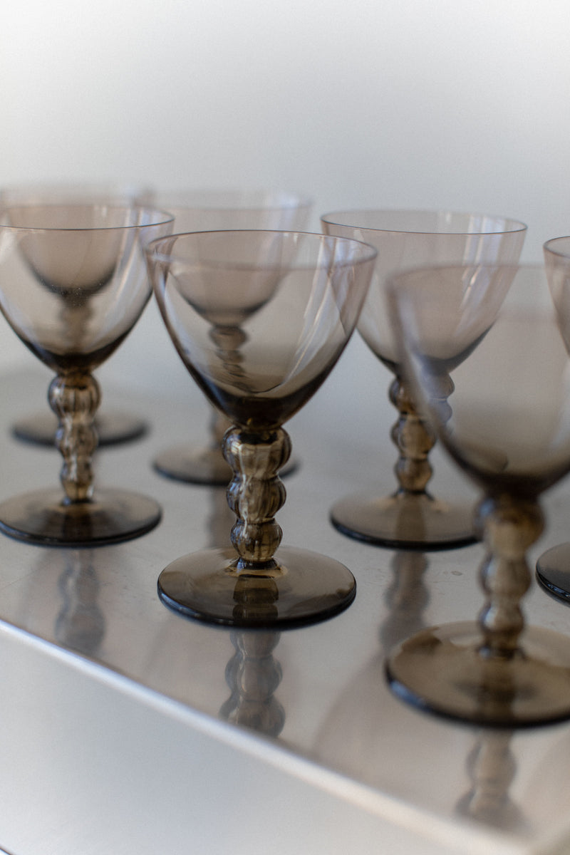 A set of Art Deco Glass goblets from Out For Lunch, featuring elegant and intricately designed stems, exudes sophistication when arranged in a row on a light surface. The focus is on the front glasses, enhancing the drinking experience as the translucent smoky gray goblets gently blur into the background.