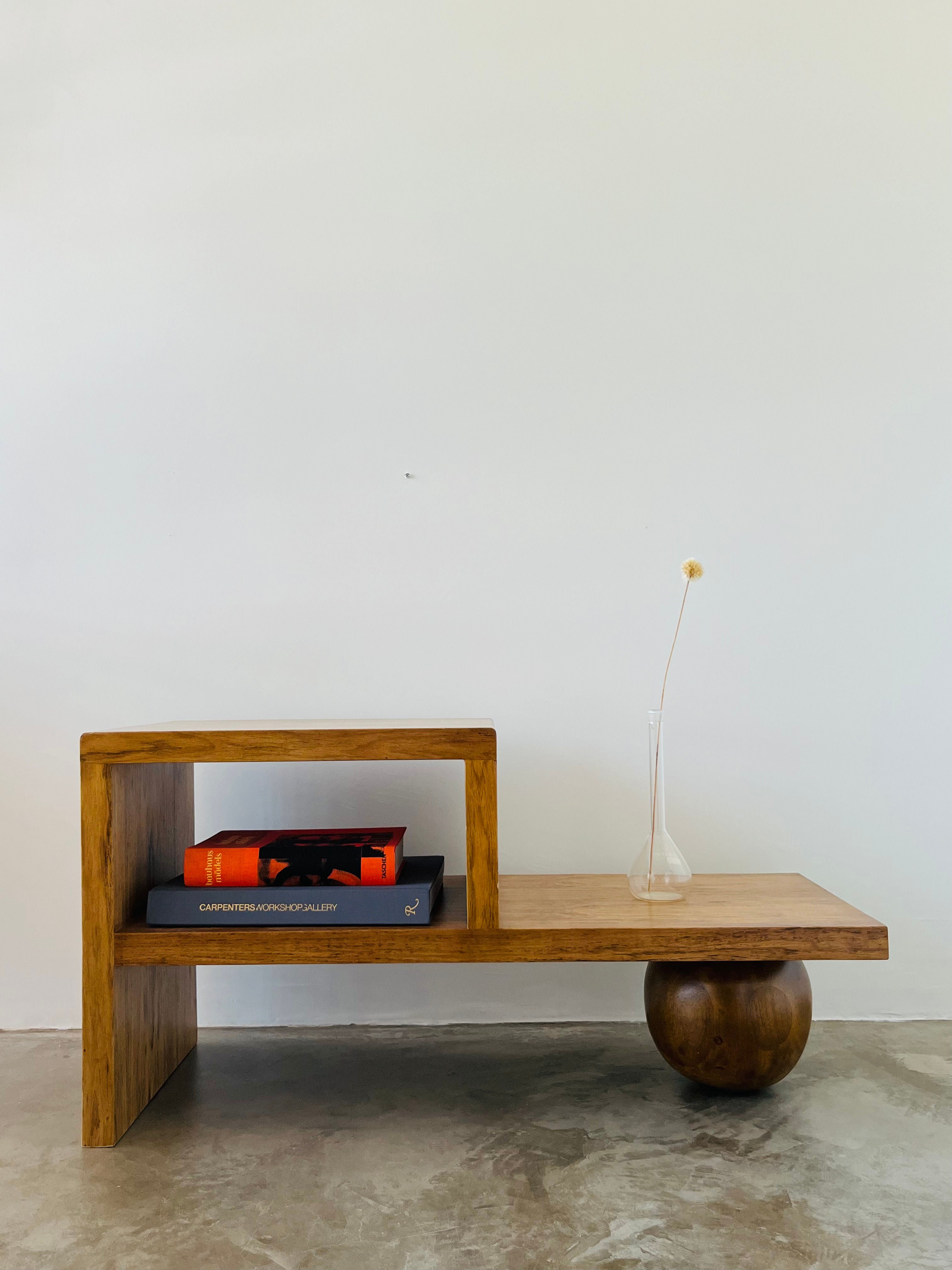 The Lina Side Table by HT., crafted from Brazilian Freijó, stands against a plain white wall with a minimalist design. It features a unique spherical base on the right and holds two books, including "CAPTIVATED," along with a small vase containing a single dried flower.