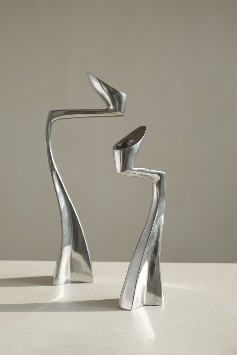 Two sleek, abstract candleholders from septembre studios stand on a flat surface. These collector’s pieces, with their elongated bodies and twisted shapes, evoke a sense of movement and balance against a plain background, captivating design enthusiasts.