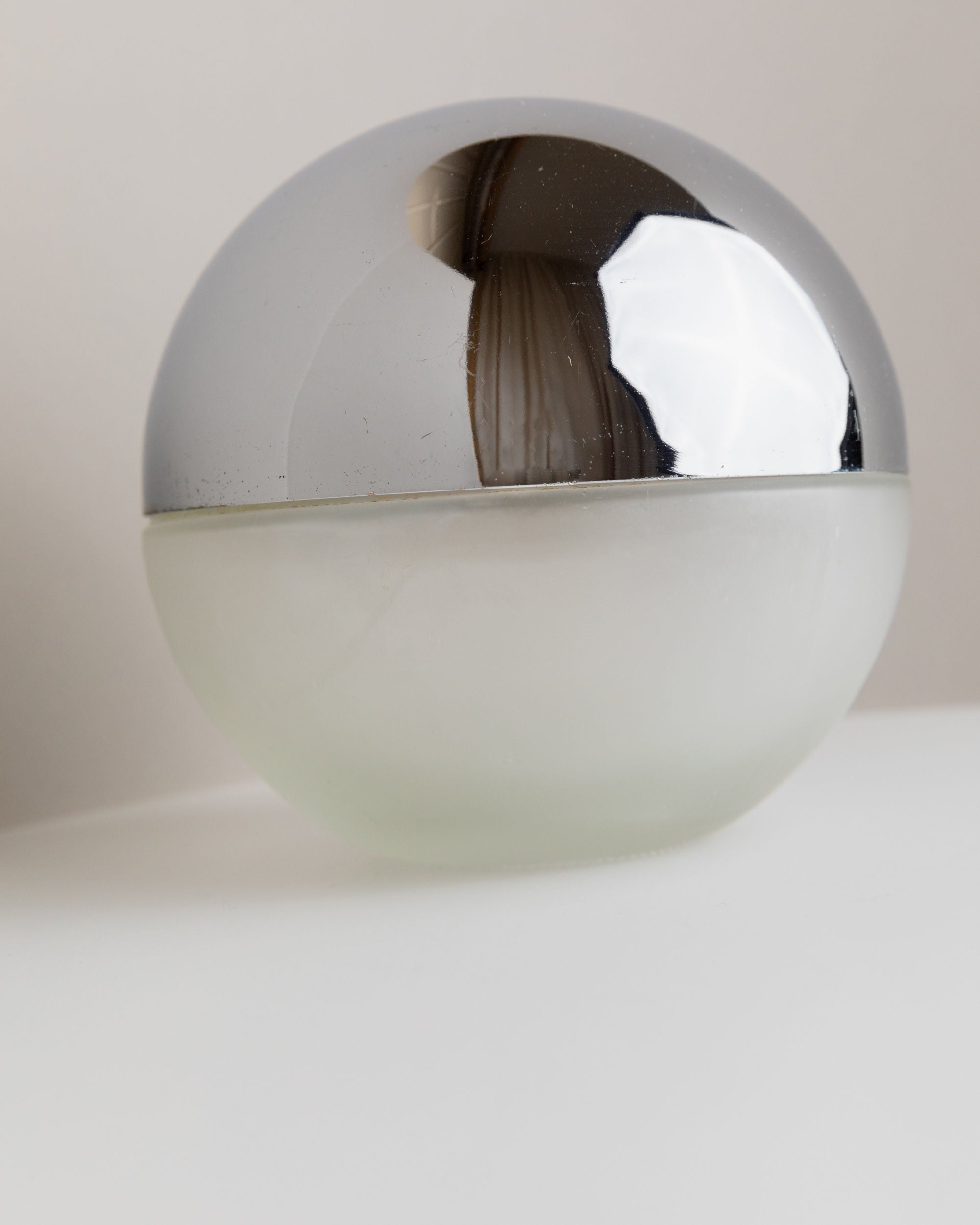 A 1980s Italian Liqueur Bottle by Bottega Jacobs, with a frosted white round base and a reflective metallic silver top, sits elegantly on a white surface against a neutral background.