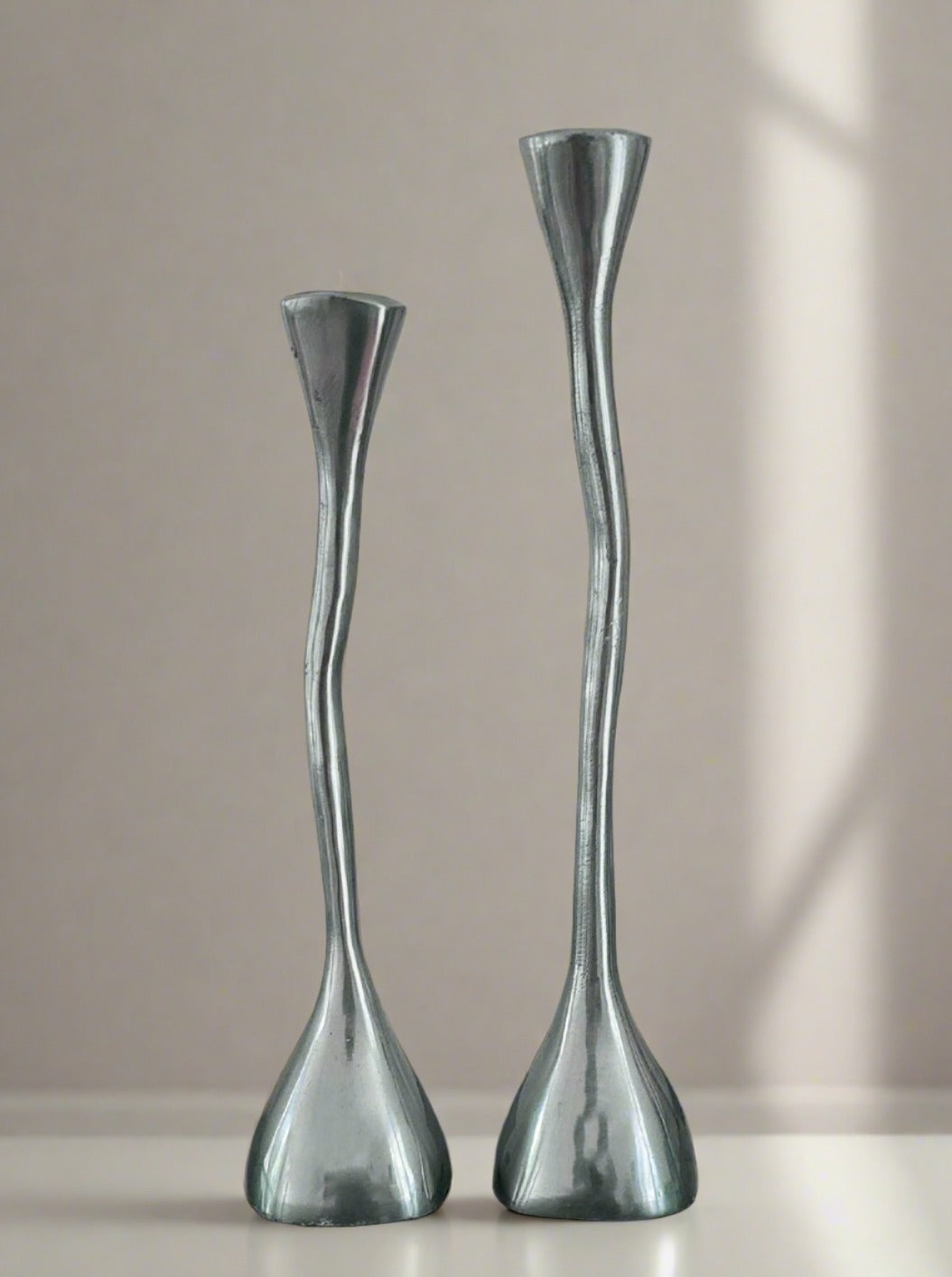 Two elegant, durable brass candlesticks of varying heights from Les Objoies' Pair of Two Candle Holders stand on a light-colored surface. Each candle holder boasts a sleek, elongated design with a subtly twisted, tapered body that broadens at the base. A soft shadow is cast against the neutral background.