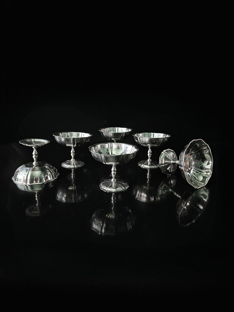 A set of seven Italian Stainless Steel Dessert Coupes by Dodo Vintage is displayed on a glossy black surface. Each dish has a reflective finish, creating mirrored reflections against a dark backdrop, highlighting the shiny texture of these elegant pieces.