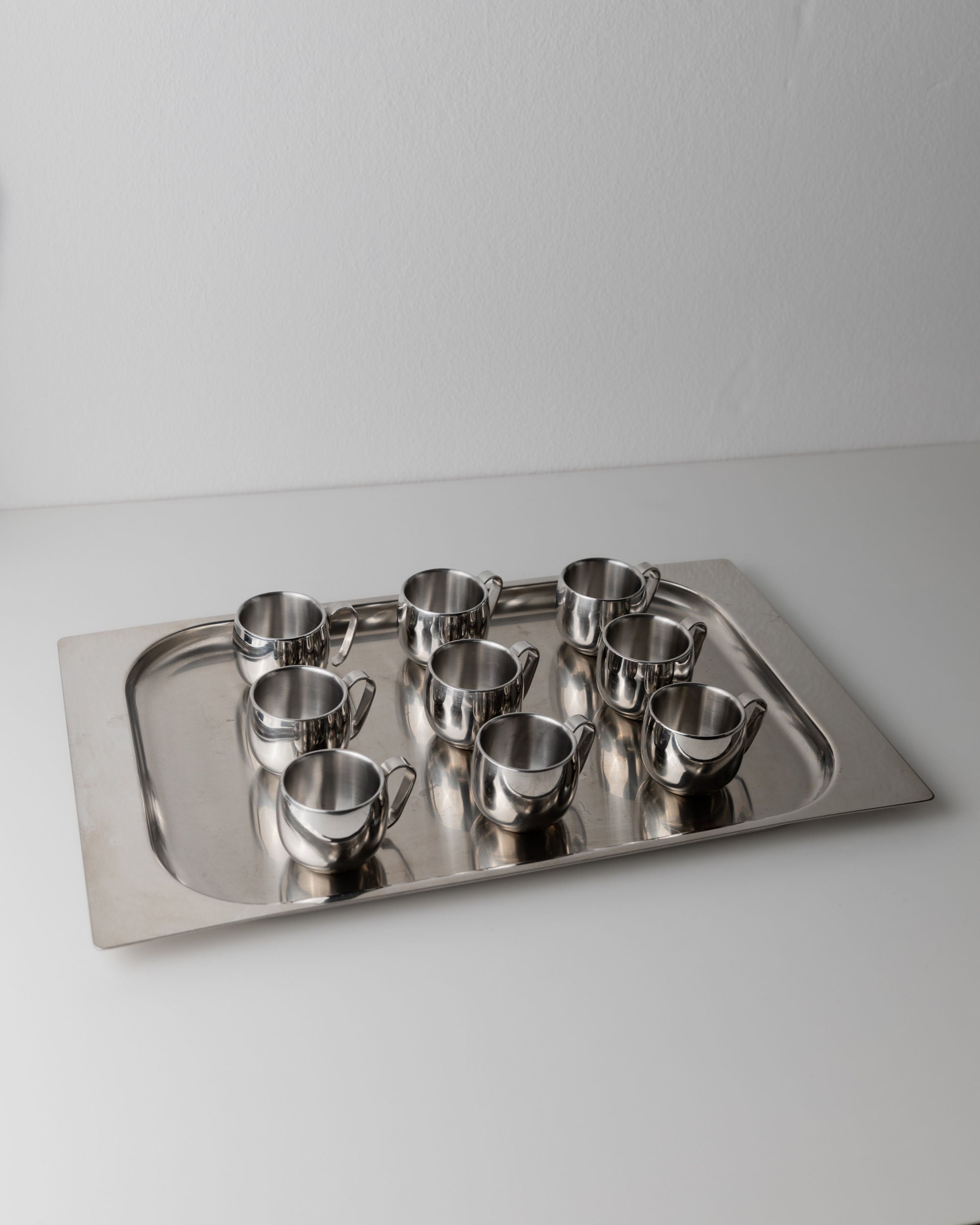 A rectangular metal tray with eight stainless steel cups neatly arranged on top, placed on a plain white surface. The Coffee set by IMCO 70s from Bottega Jacobs combines vintage charm with minimalist elegance and functionality.