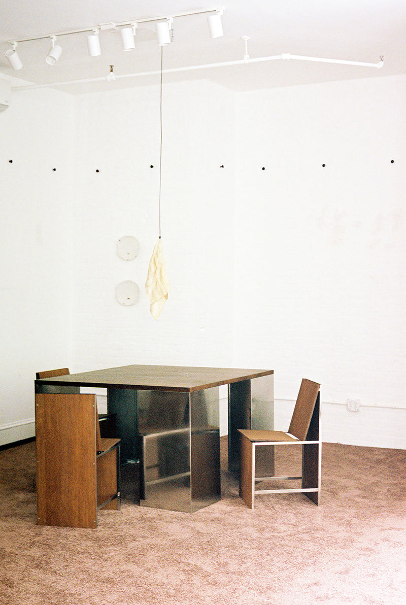 A minimalist room with a Square Table 2022 by Studio HAOS, three wooden chairs, and metal legs exudes grounded elegance. A beige-shaded light hangs above, while white walls and a brown carpet complete the serene space.