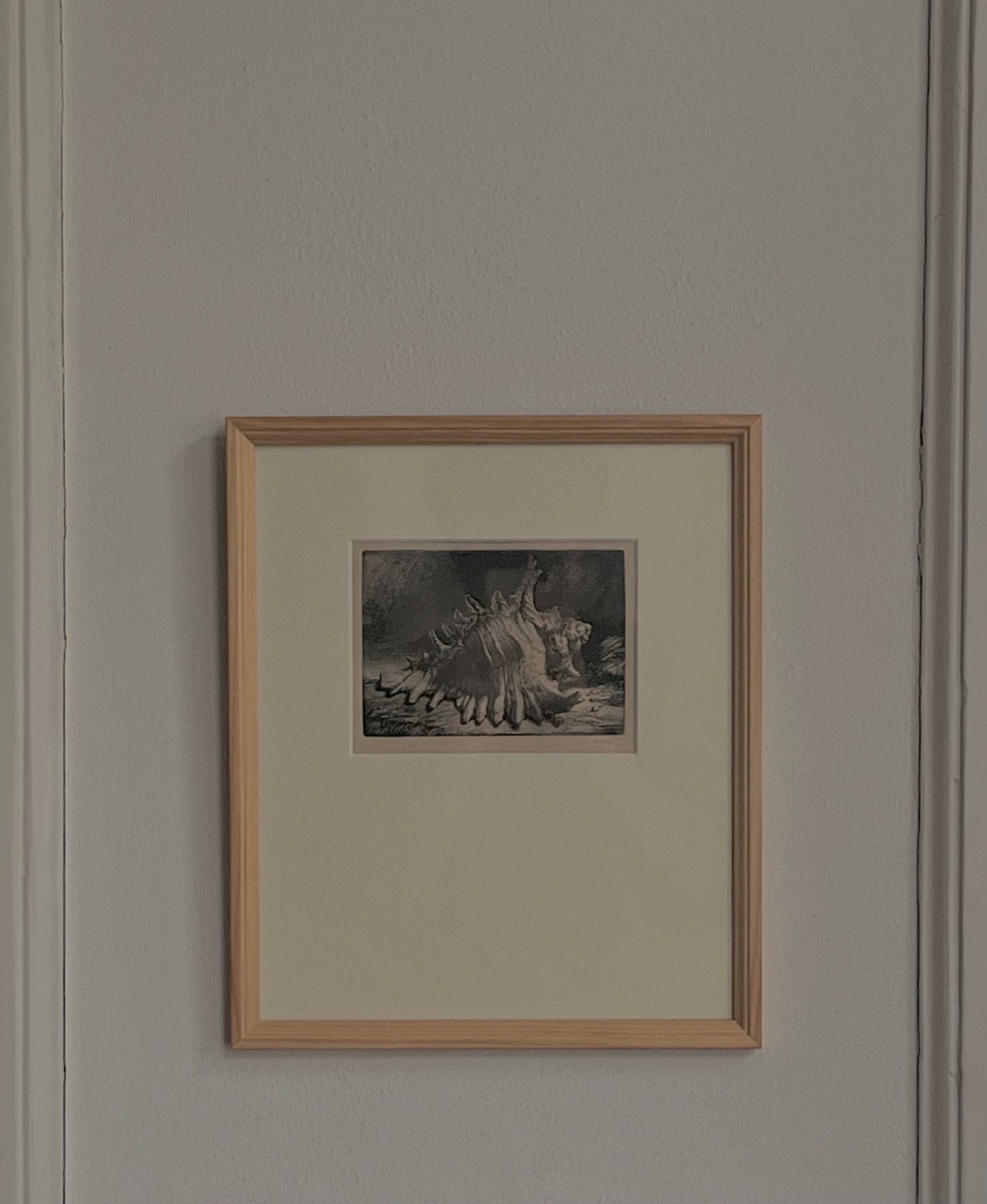 A framed artwork, "Shell Etching" by Médecine, hangs on a light gray wall. It features a natural oak frame and showcases a surreal, abstract scene with muted colors and delicate details by the Swedish artist.