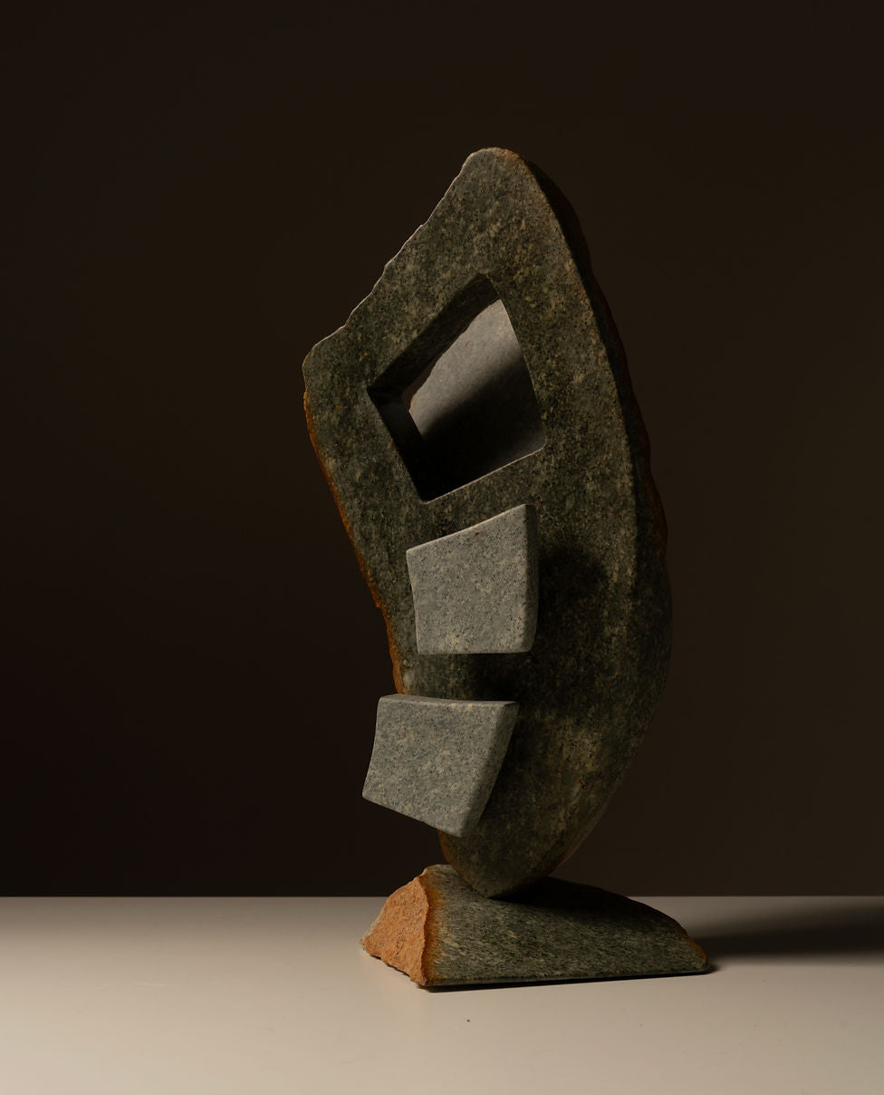 The "Two-toned Abstract Sculpture" by Les Objets Amsterdam is a handmade piece crafted from natural stone with an irregular shape and three vertically arranged rectangular cutouts. Its textured surface features a blend of dark and light tones, embodying modernist art sensibilities.