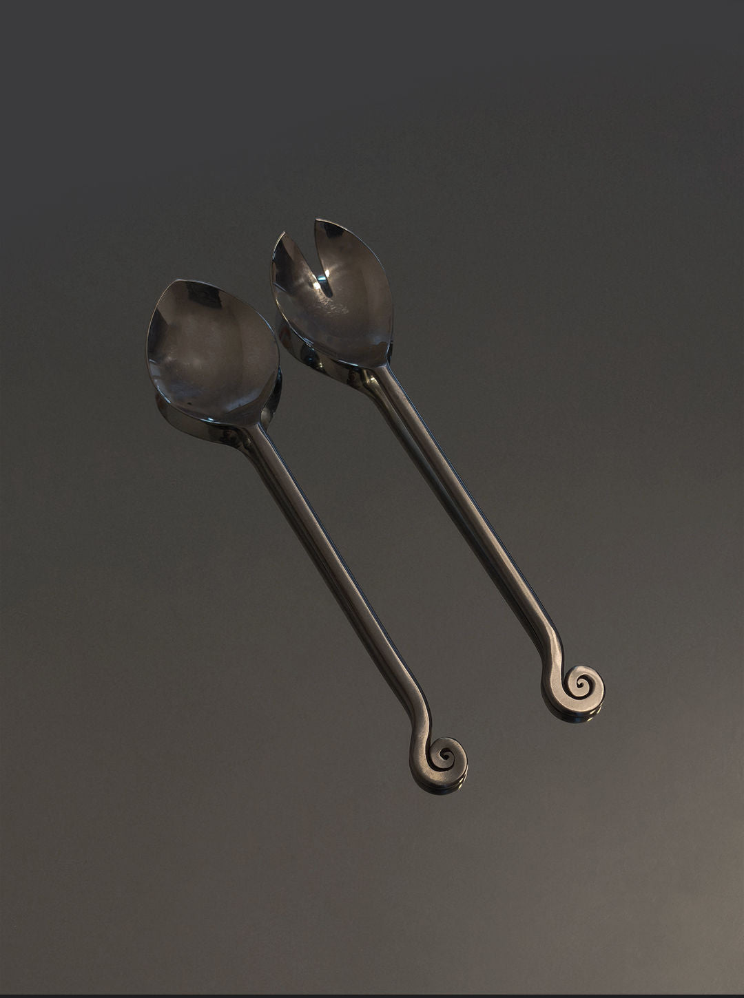 Les Objoies Spiral Salad Servers feature a polished brass spoon and fork with long handles ending in spiral designs, adding decorative charm when displayed together on a dark surface.