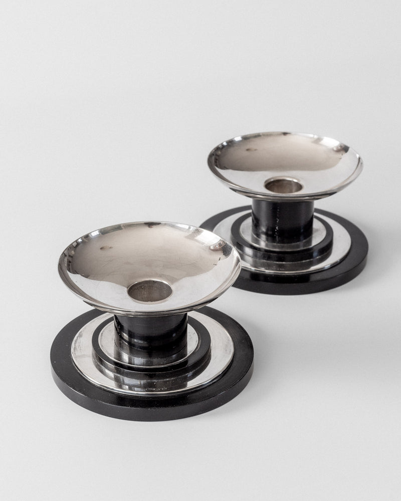 Two round Spigel Modernist Sterling Silver Candleholders (1934) with wide, shallow bowls and circular bases sit on a plain light background. Their geometric black and silver design offers a sleek, contemporary look.