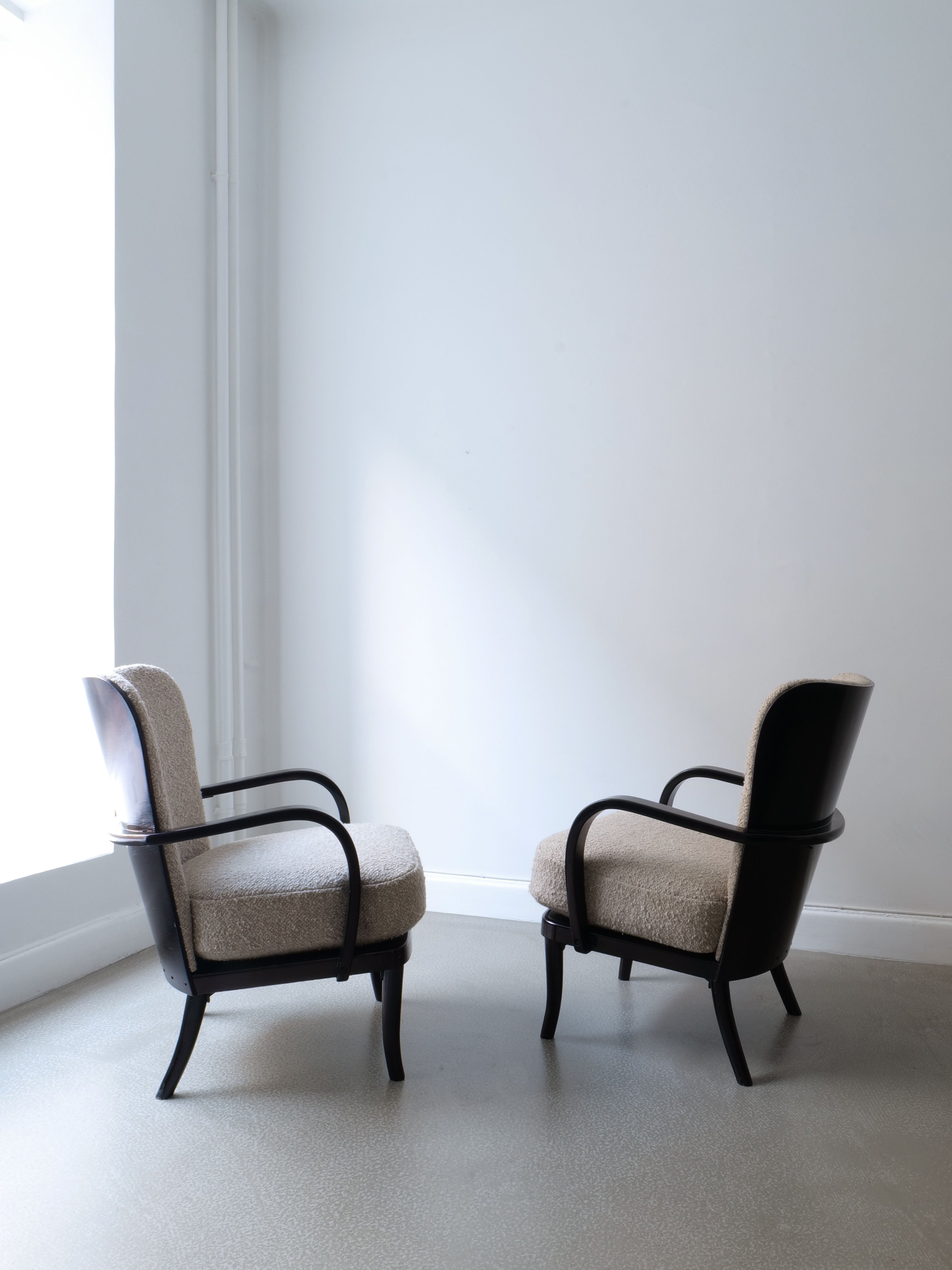 A pair of Werner West Armchairs from the 1930s by Collection apart are positioned facing each other in a minimalist room, featuring black lacquered wood frames and Moretti bouclé upholstery. The space has a smooth gray floor and a white wall, with natural light streaming through a large window.