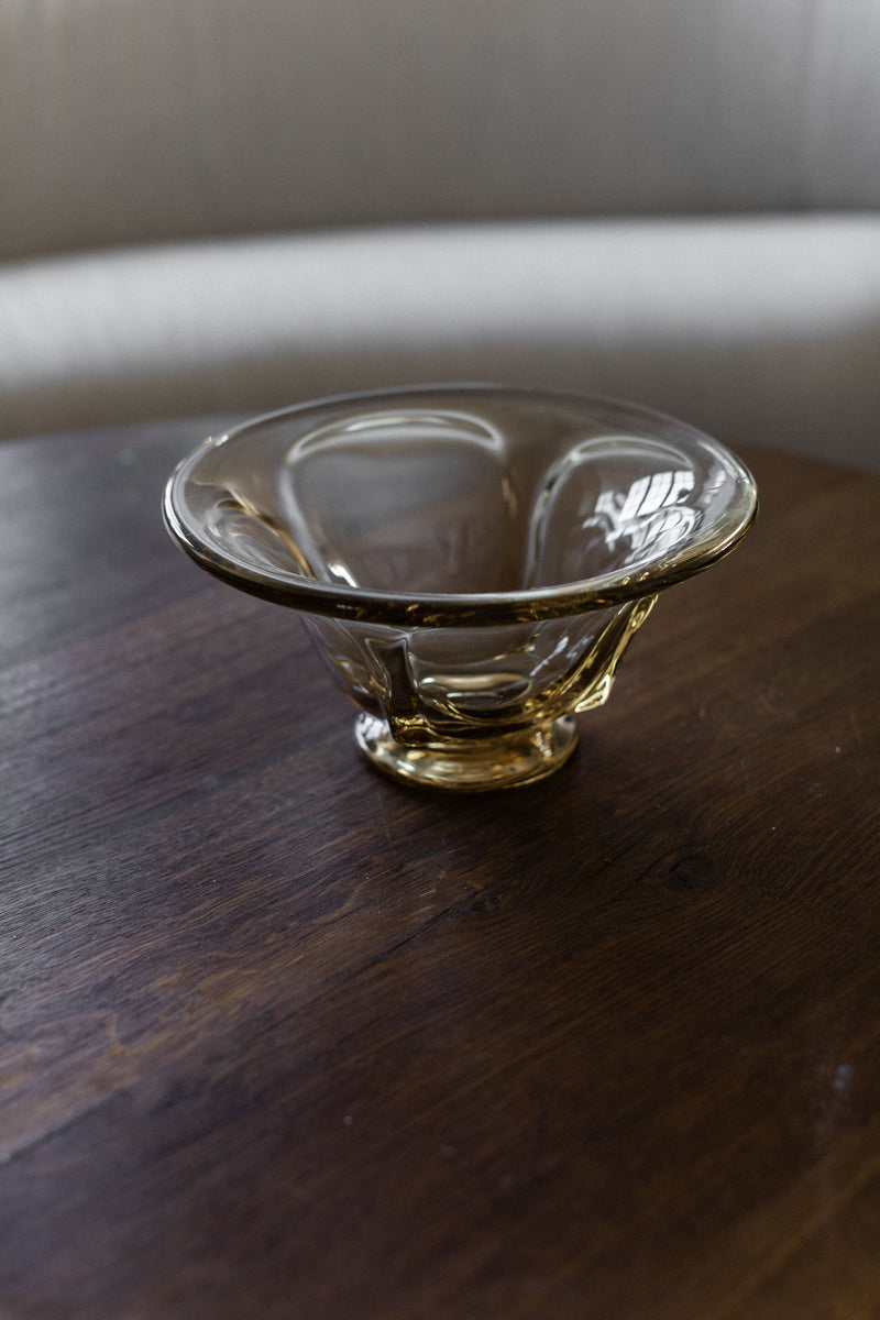 A Vintage Glass Bowl by Out For Lunch rests on a dark wooden table, its mid-century design accentuated by the room's gentle lighting. The warm glow dances across its surface, with a blurred sofa subtly appearing in the background.