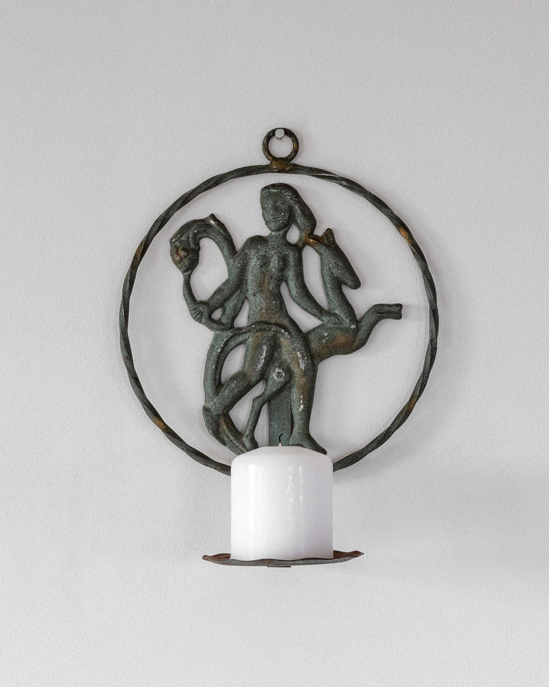 This Spigel Art Deco wrought iron wall sconce from the 1920s/30s features a circular frame with a figure entwined with a naga. It holds a white pillar candle, casting warm light against the light gray wall behind.