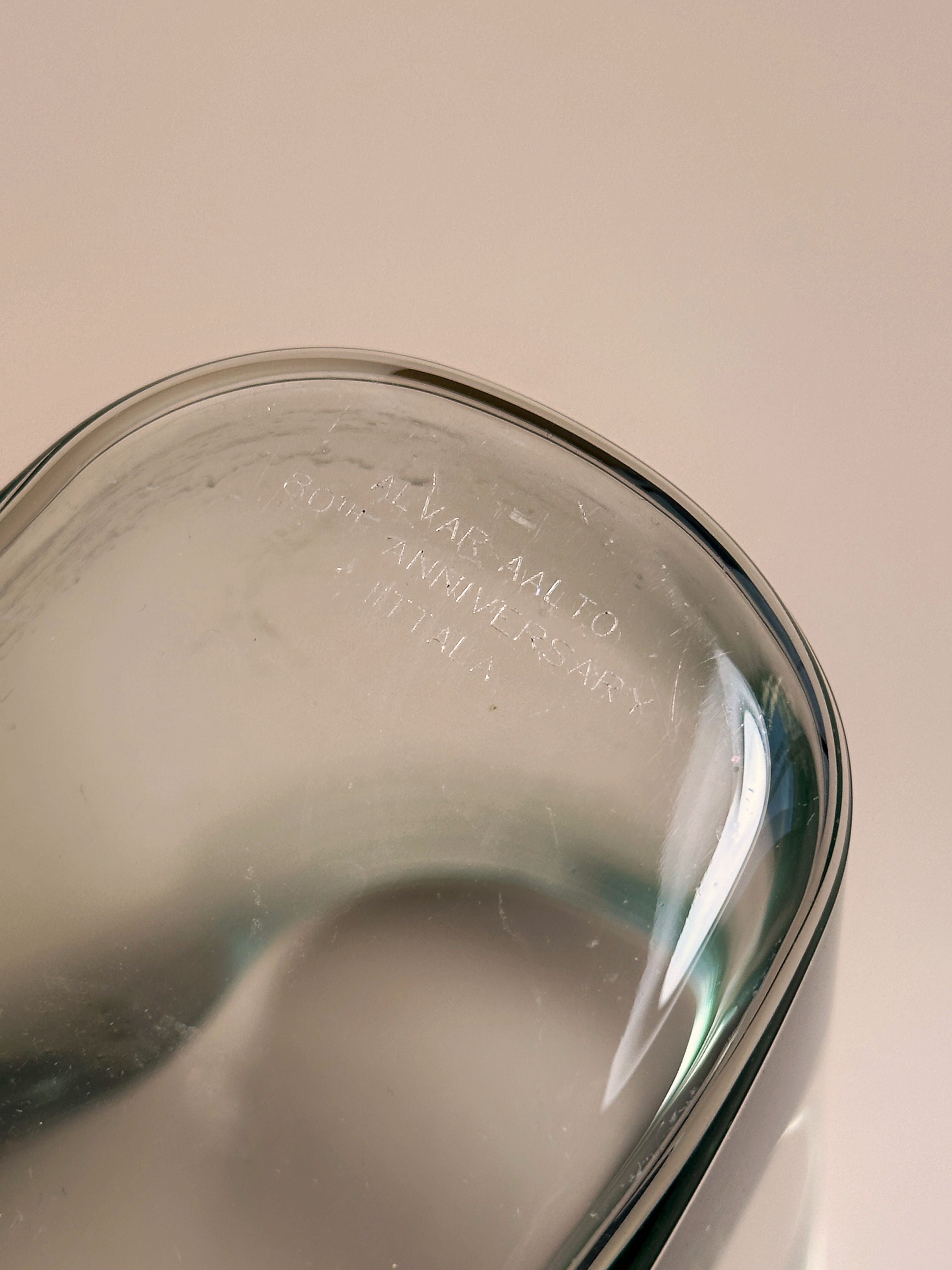 Close-up of the base of a transparent glass object with engraved text that reads, "Alvar Aalto Collection 90th Anniversary Iittala." This Limited Edition Aalto Vase from Dodo Vintage has a smooth, curved shape and a slight greenish tint, embodying the iconic design of an Aalto Vase.