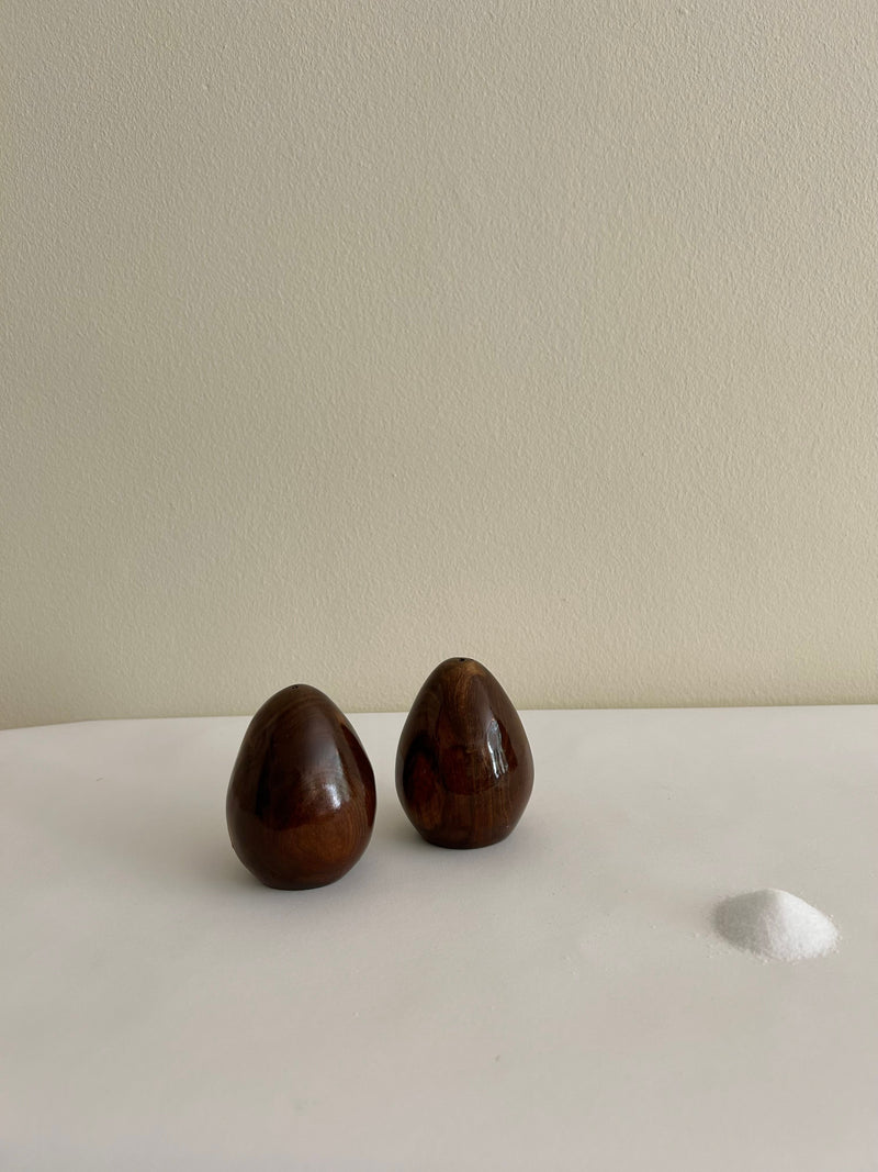 The Vintage Danish Wooden Salt & Pepper Set from Flock Studio, featuring two polished dark brown wooden eggs, is displayed like treasures in a vintage tableware collection on a white surface next to a small pile of white sand, set against a neutral light-colored wall.