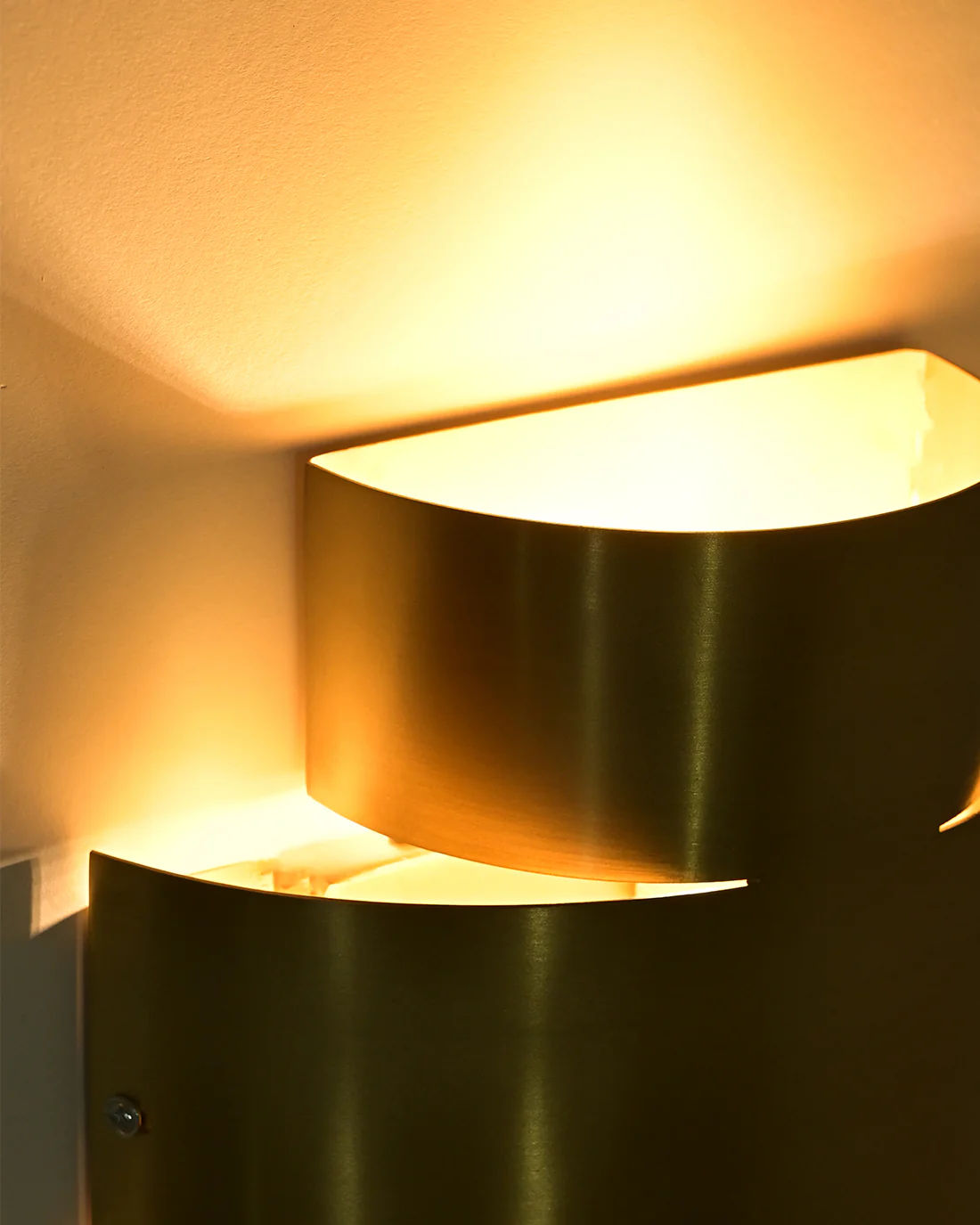 The Aura Wall Sconce by LMNOH features a sleek metallic cylindrical design that emits a warm, soft glow. This brass sconce creates ambient patterns on your wall, enhancing the cozy atmosphere of contemporary interiors.