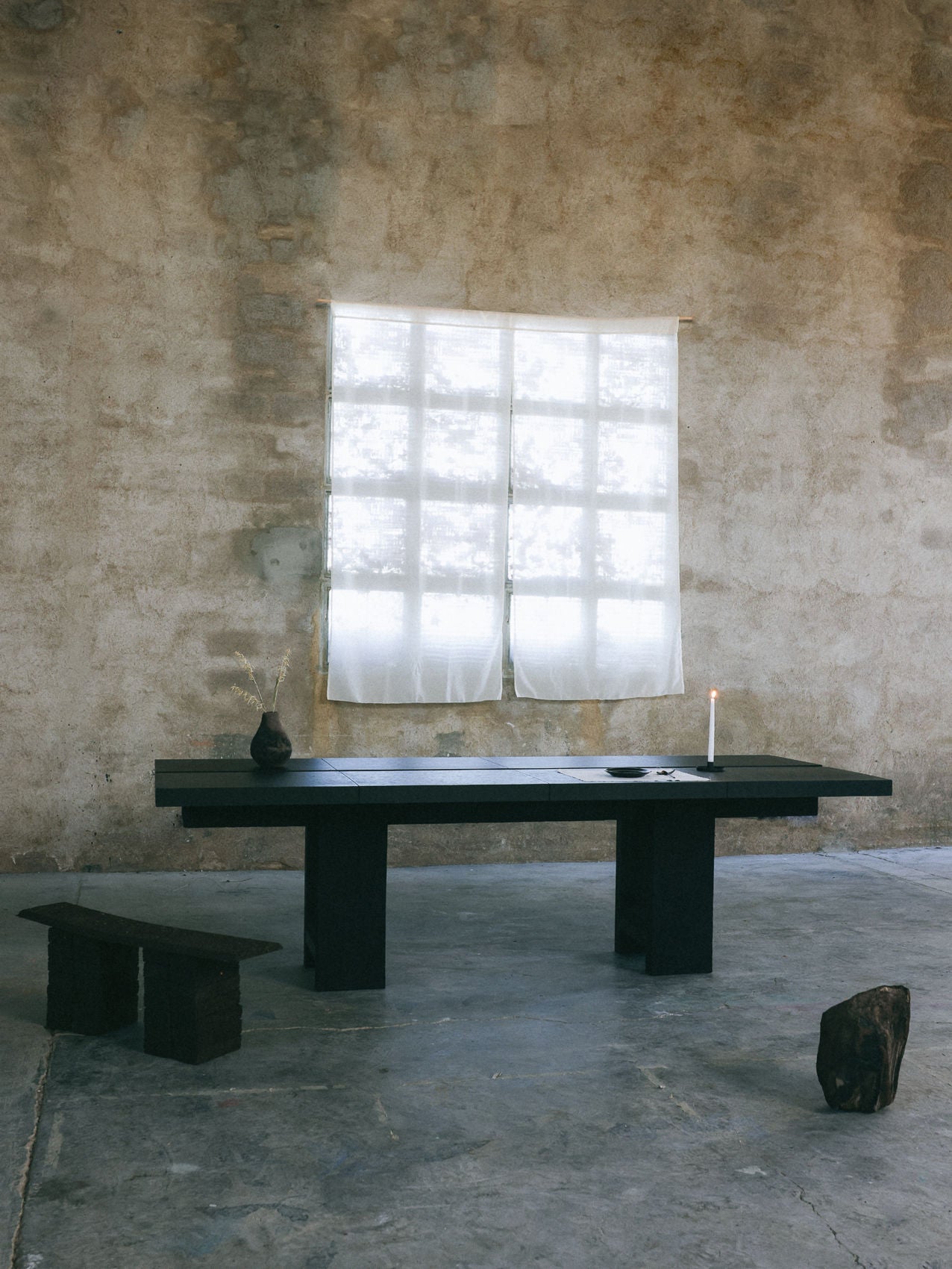 A minimalist room with a textured stone wall and a large window draped in sheer curtains features the Twin Oaks Dining Table by Obstacles. The table holds a lit candle and vase, while a small bench, wooden block, and stone enhance the space's rustic yet uncluttered vibe.