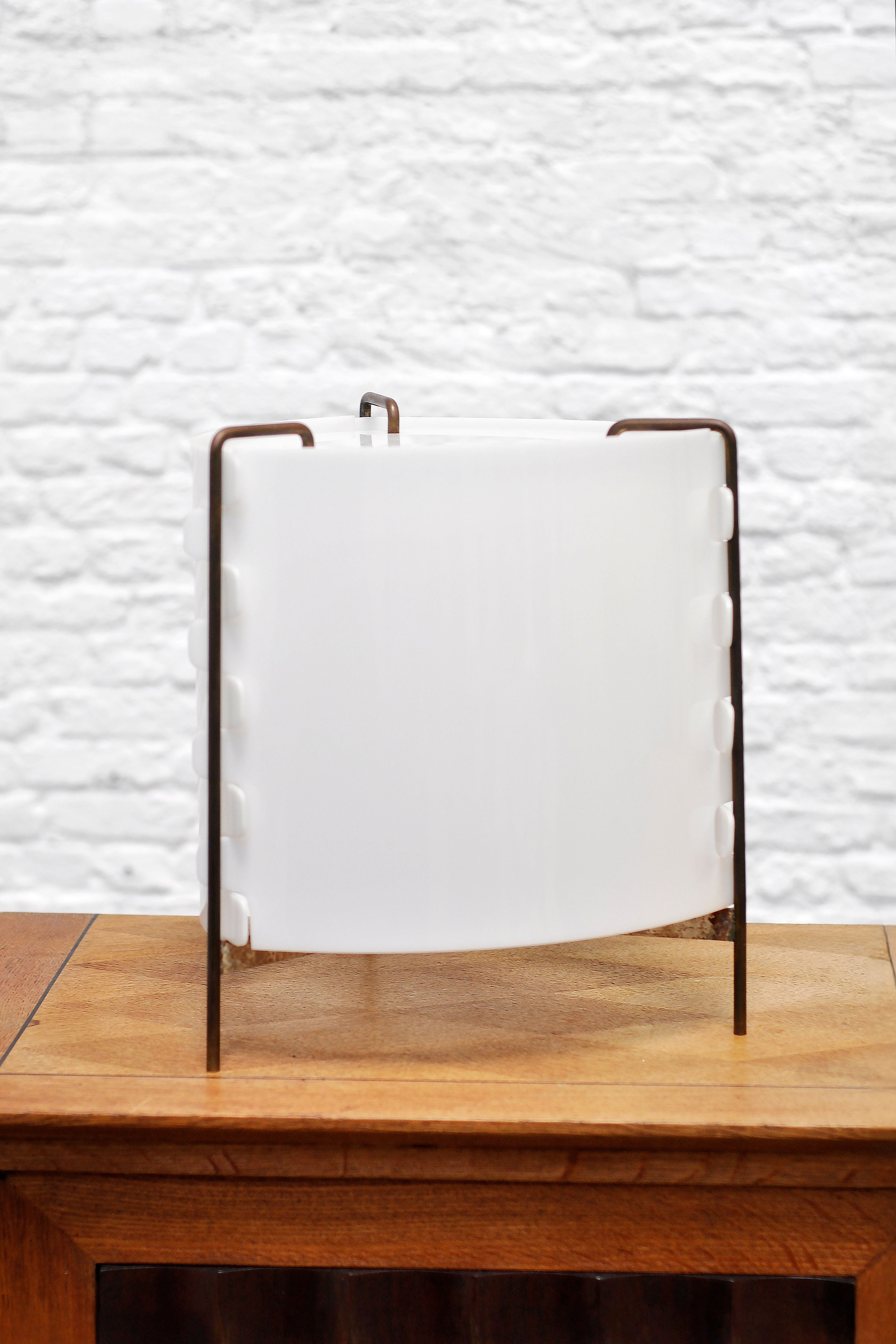 A semi-translucent white panel mimicking the 'M8 Fanal' lamp by Joseph-André Motte for Huchers-Minvielle (France, 1958) rests on a wooden table with a black frame, set against a white brick wall, exuding French mid-century modern elegance. Brand: Introverso.