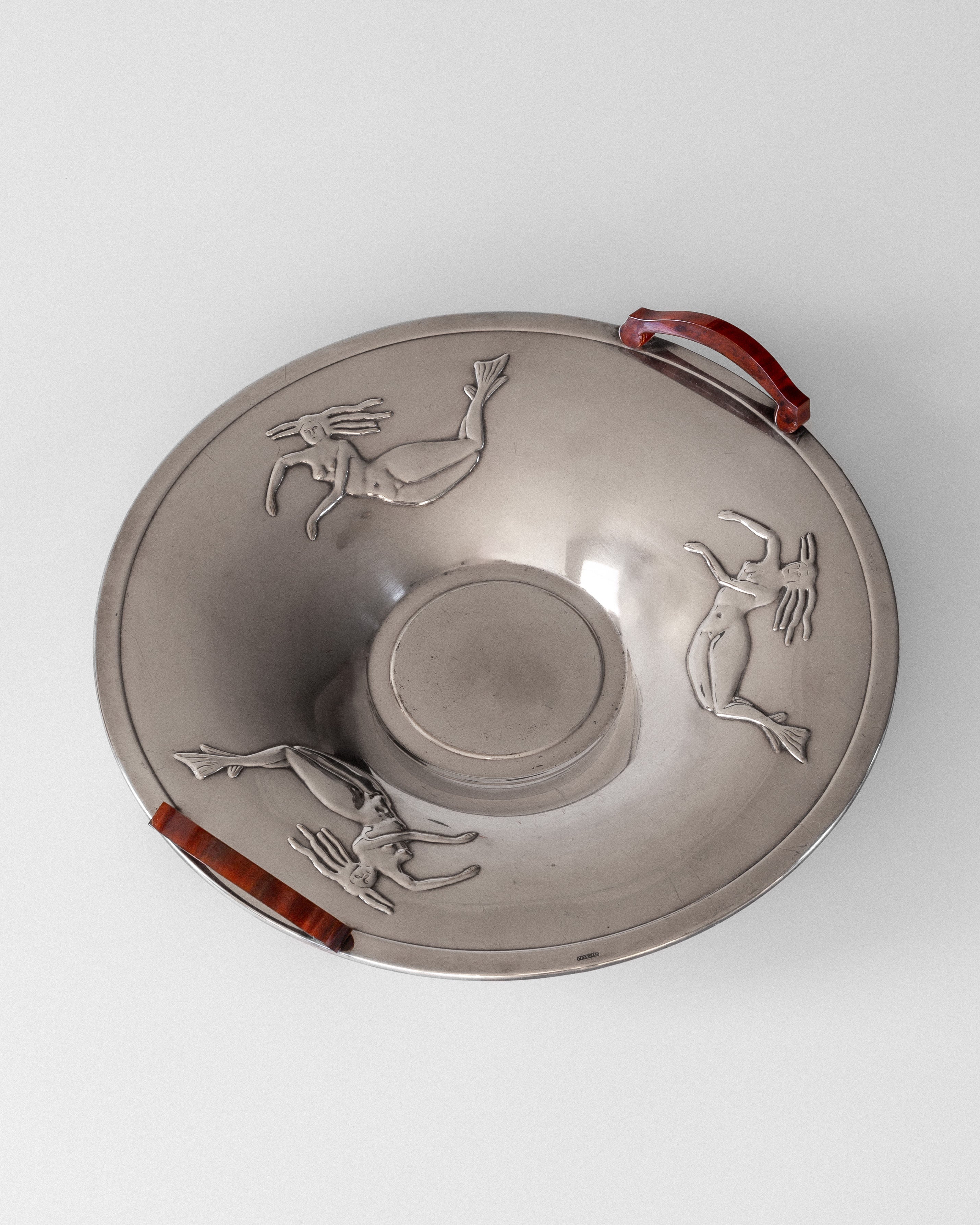 A Swedish 1930s Spigel silver-plated dish features a round shape with a raised center, two red Bakelite handles, and embossed Art Deco figures. It is displayed against a plain white background.