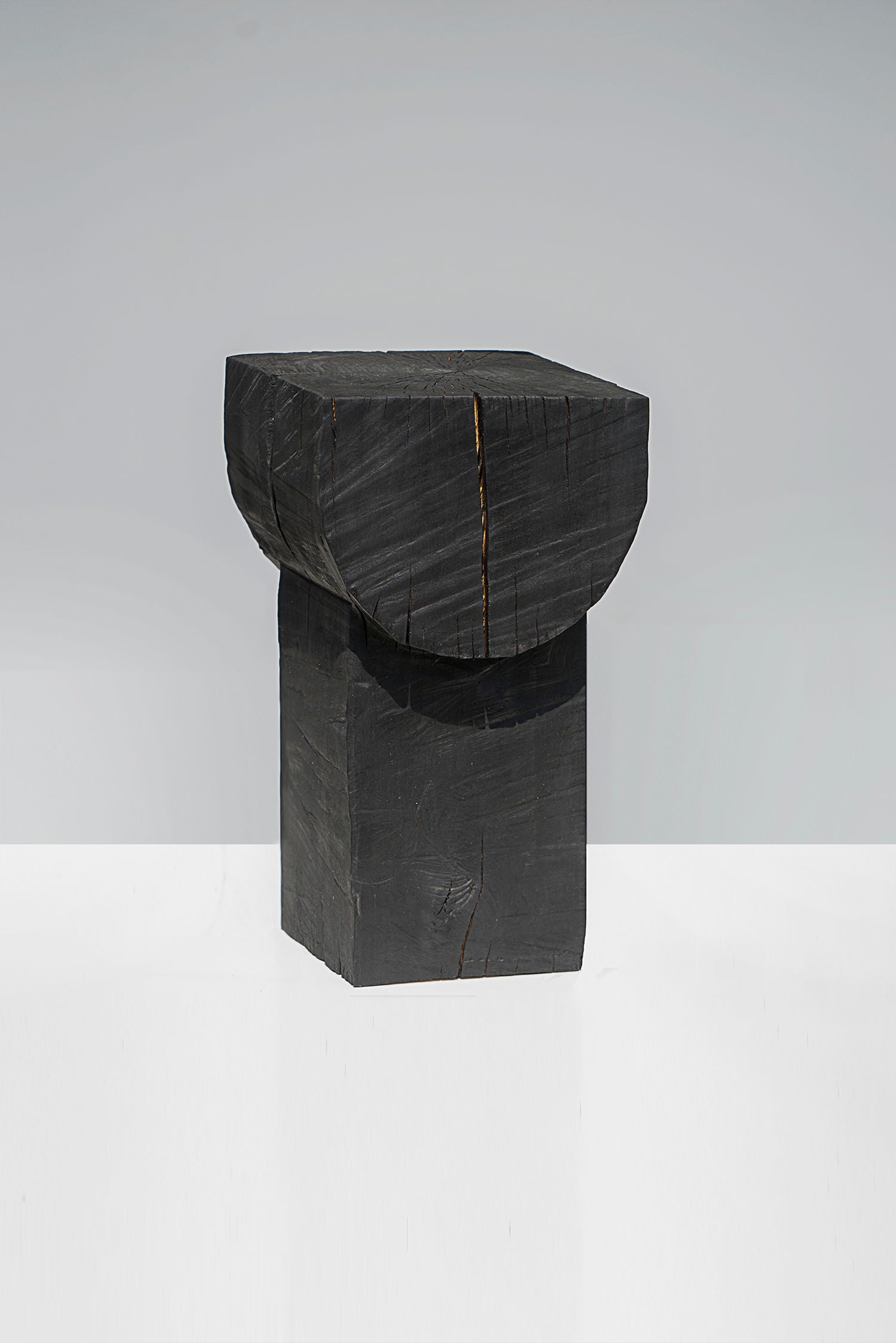 The "Burnt Black Coffee Tables/Stools" by Touch With Eyes is a modern wooden stool with a geometric design, featuring a dark square base and rounded flat top. Its handmade finish reveals the deep charcoal-stained wood grain, set against a plain gray backdrop.