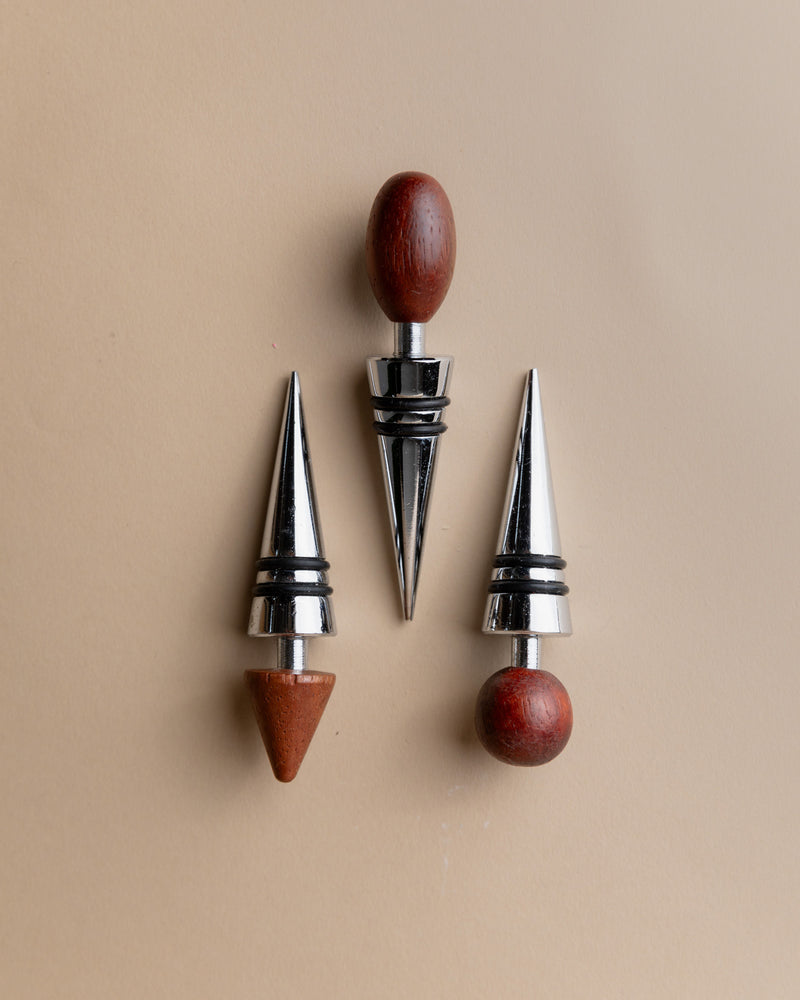 Three Bottega Jacobs 3 Bottle Stoppers, Mid Century Italy 1980s, feature round and cone-shaped wooden tops with shiny silver bases and black rings, elegantly displayed on a beige surface—perfect vintage wine accessories.