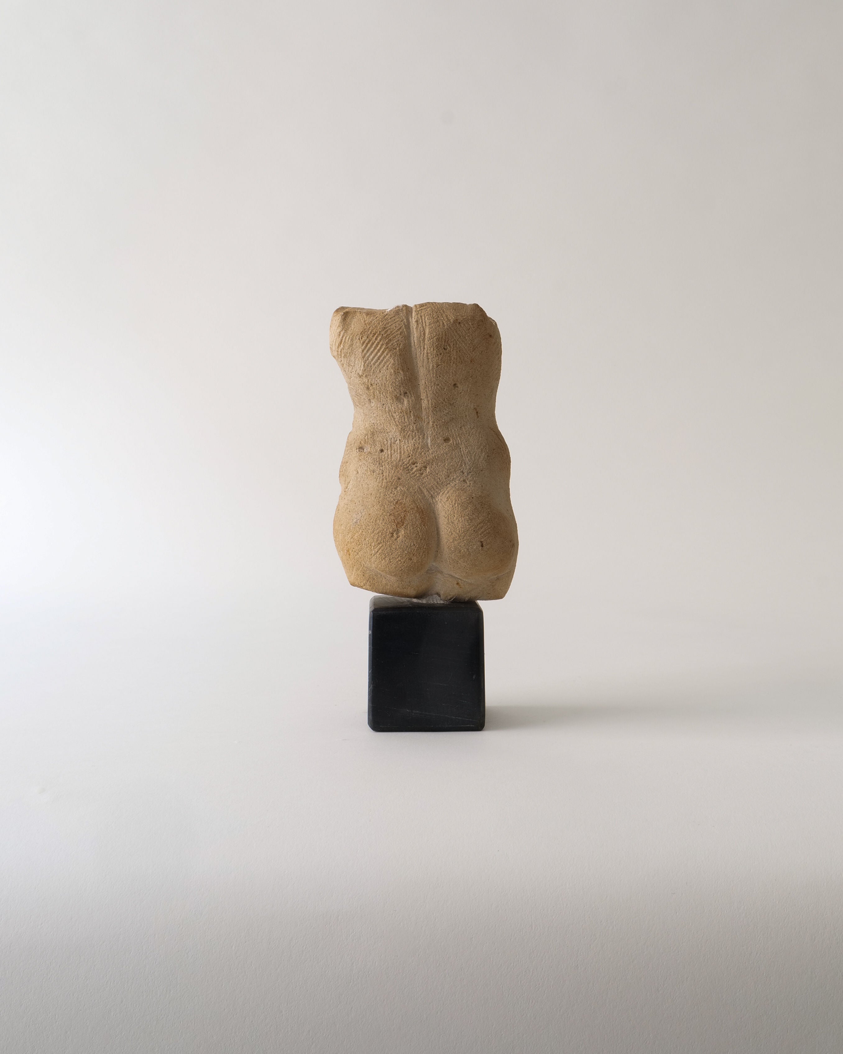 The Torso Sculpture by Misja Kristoffer Rasmussen (b.1971), from Collection apart, is a small beige stone piece inspired by classical sculpture, showing a headless, armless torso on a black rectangular base against a light background.