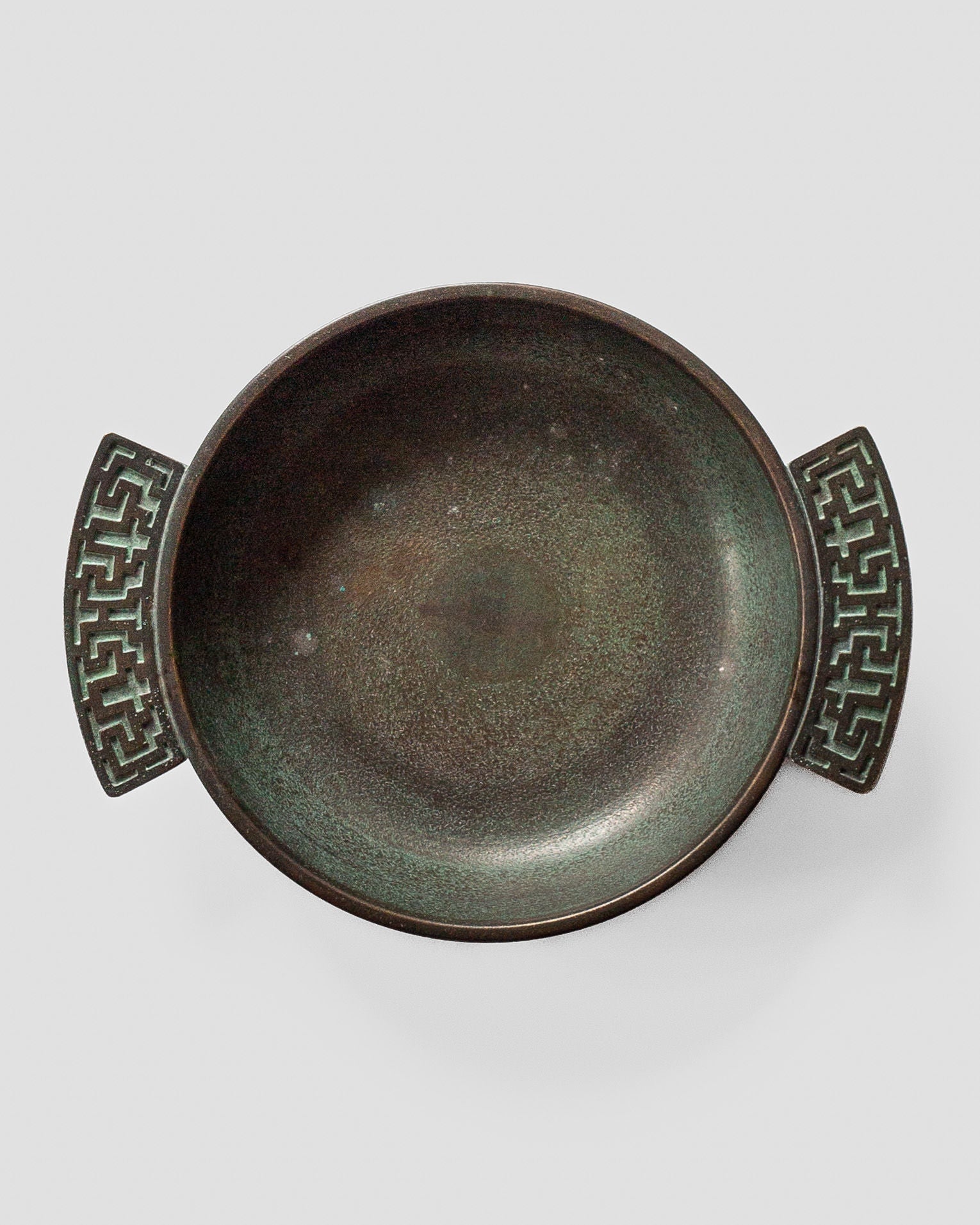 A round Art Deco bronze bowl from the 1930s by Spigel, featuring Greek key handles with geometric cut-outs and a worn surface with greenish patina.