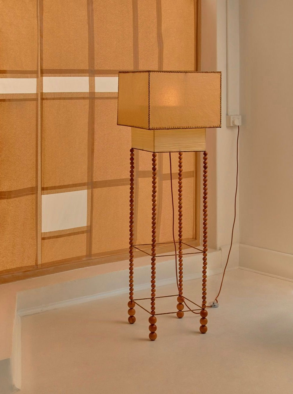 The "Feels Like Home" Floor Lamp by Lana Launay features a modern, rectangular lampshade atop a tall, decorative wooden stand with beaded legs. This hand-made lamp is placed in a minimalistic room with light-colored walls and a large window draped with a sheer beige curtain. An electrical cord is visible on the right side, contributing to the soft ambiance.