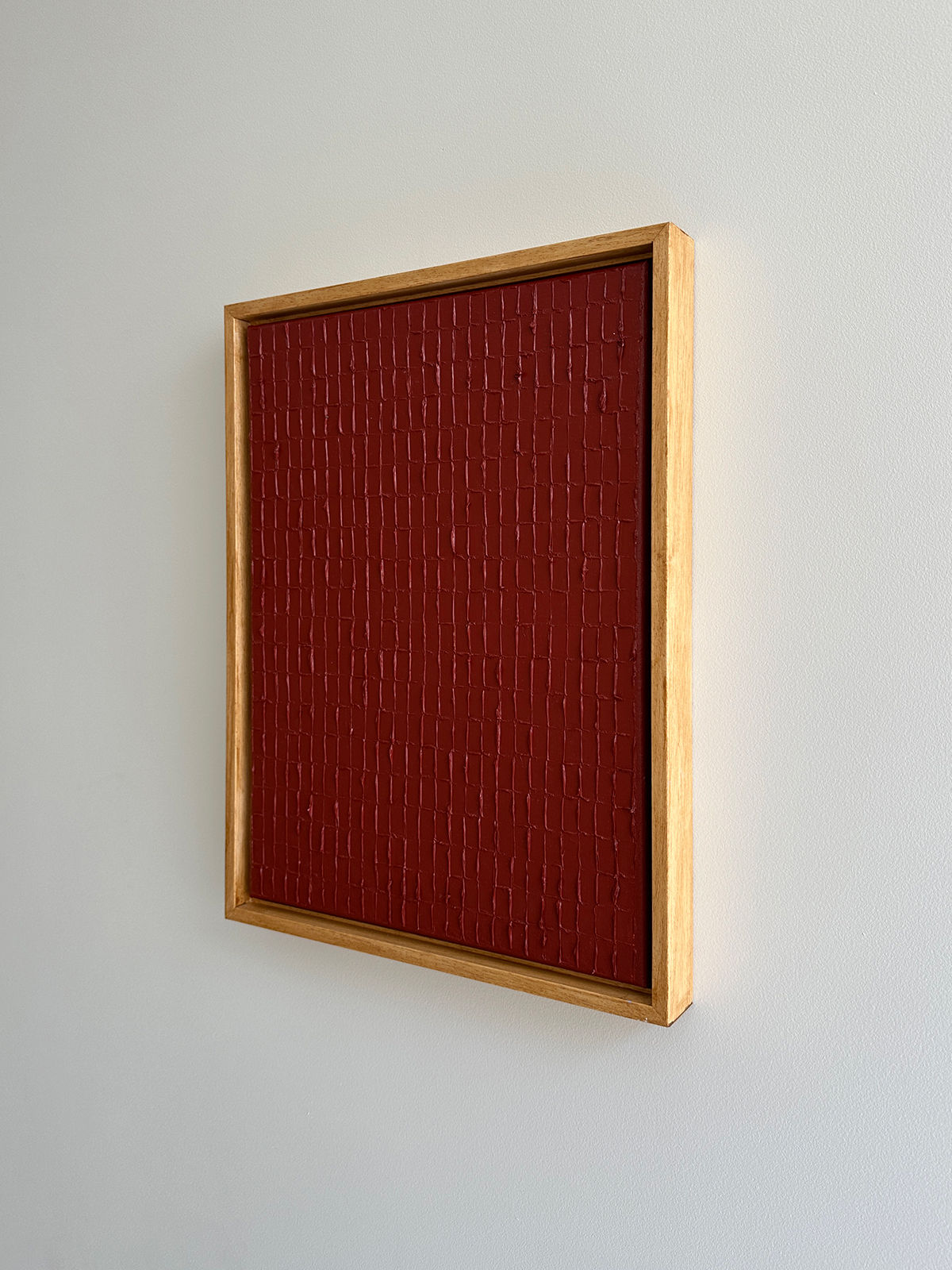 Framed artwork hanging on a white wall, featuring a textured, deep red surface with a grid-like pattern. The frame is simple and wooden, contrasting with the bold color and design inspired by Korean Dansaekhwa, inviting mindfulness through its intricate yet organic forms. This piece is titled "Indian Red - Oil on canvas" by Roi Elmaliah.