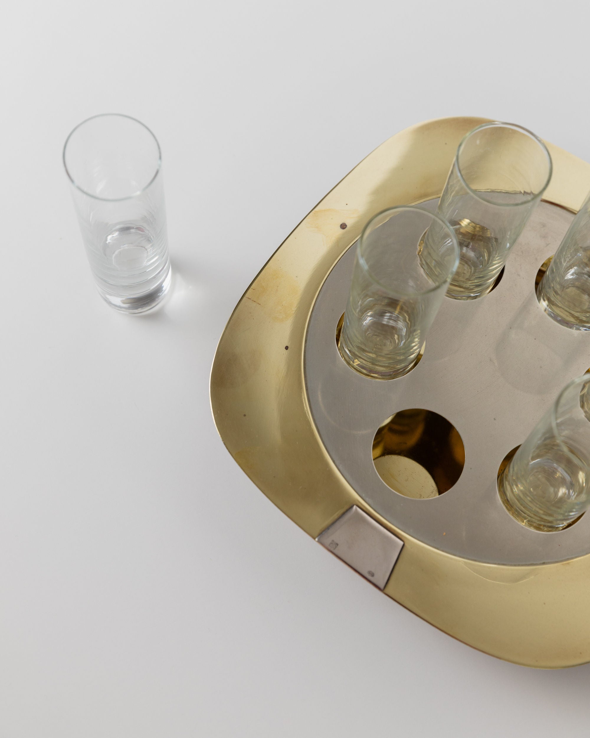 On a white surface, the Bottega Jacobs "Set Shot Glasses and Tray - Mid Century Design 70s" features four small vintage shot glasses on a gold-colored tray with mid-century design elements, plus an identical empty glass nearby, serving as a chic decorative accent.