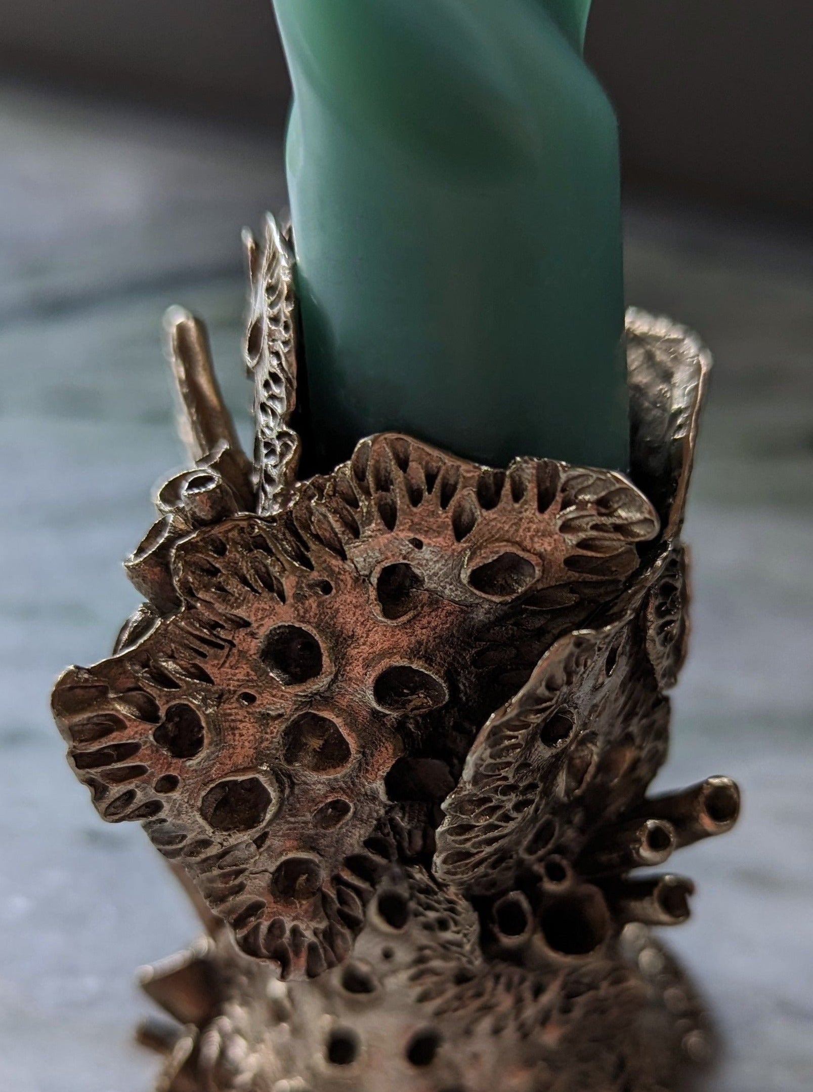 A close-up image of a Sophie Symes Bronze Lichen Candle Holder crafted from metal, designed with intricate patterns resembling lotus seed pods that encircle a teal candle.