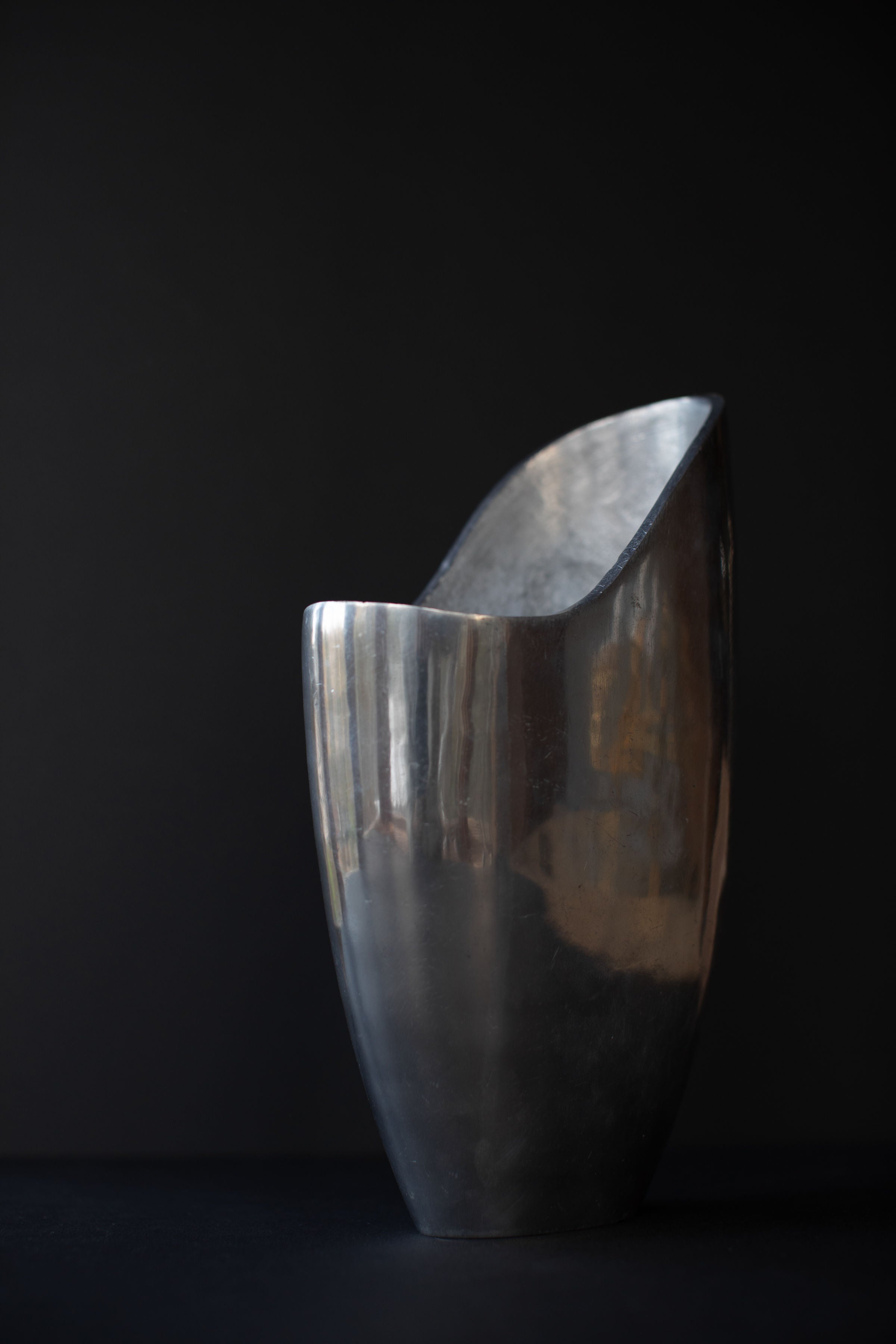 The Aluminum Cast Vase by Out For Lunch is a polished, abstract metallic sculpture with a smooth, reflective surface against a dark background. Its tall, curved form gleams with light reflections, exuding an elegant and minimalist presence that embodies an industrial aesthetic.