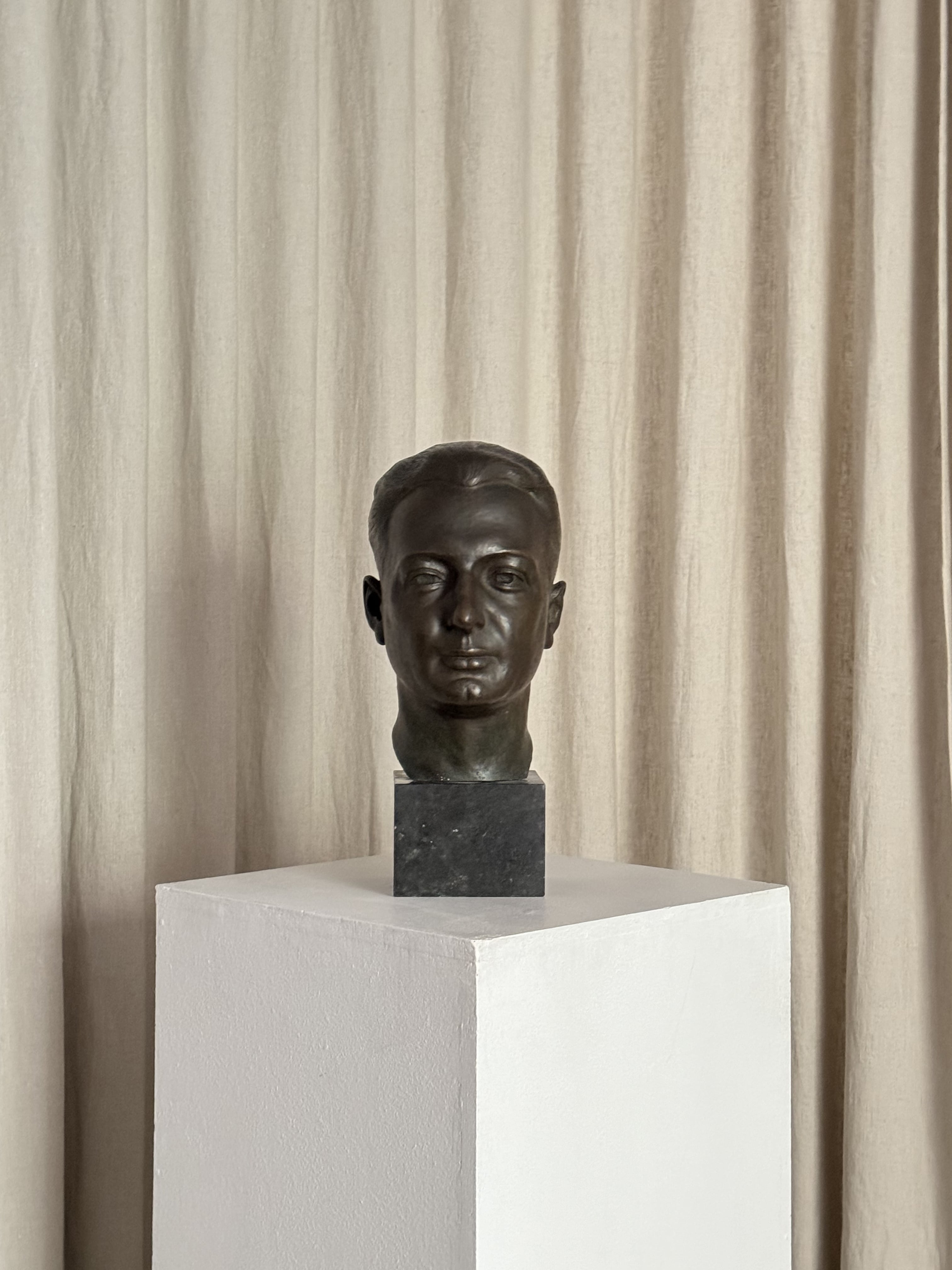 The "Testa Virile" Sculpture 1930s by FEBRERO SHOP, a bronze bust displayed on a white pedestal before beige curtains, features a realistic head and neck, creating an elegant and minimalist atmosphere.