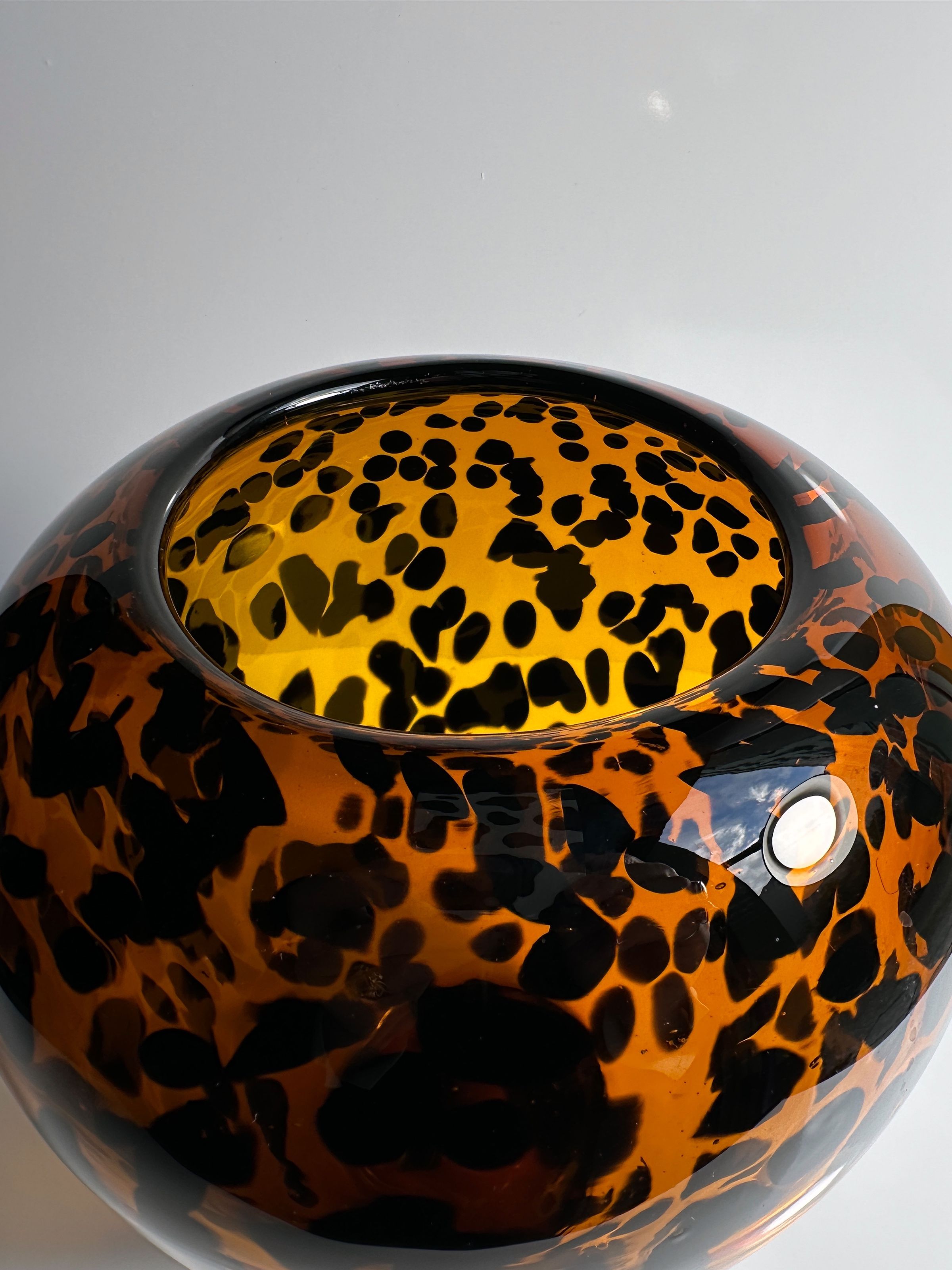 This spherical glass vase, known as the "Tortoise vase Murano for Christian Dior - 1960s" by Dodo Vintage, boasts a vibrant leopard print pattern in orange, black, and yellow. Its glossy surface and open top reflect light beautifully, reminiscent of classic Murano Tortoise designs.